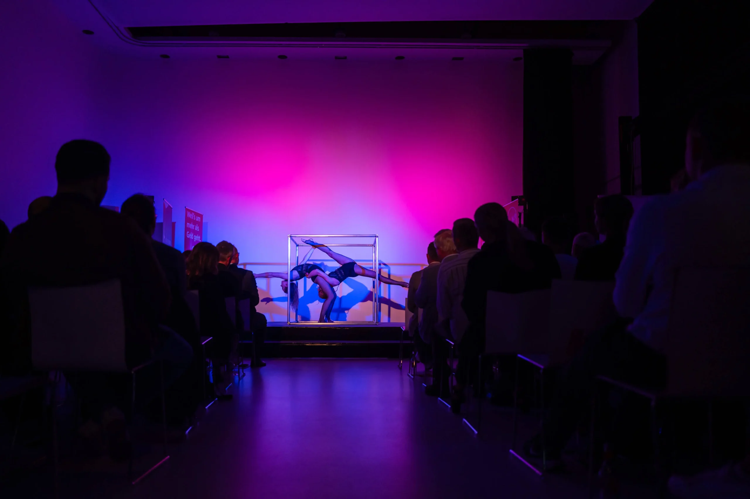 Während einer Duo Acrobatic Cube Show krümmt sich ein Artist in einem Glaskasten auf der Bühne, der von rosa und violetten Lichtern beleuchtet wird, während das Publikum in einem abgedunkelten Theater sitzt und zusieht.