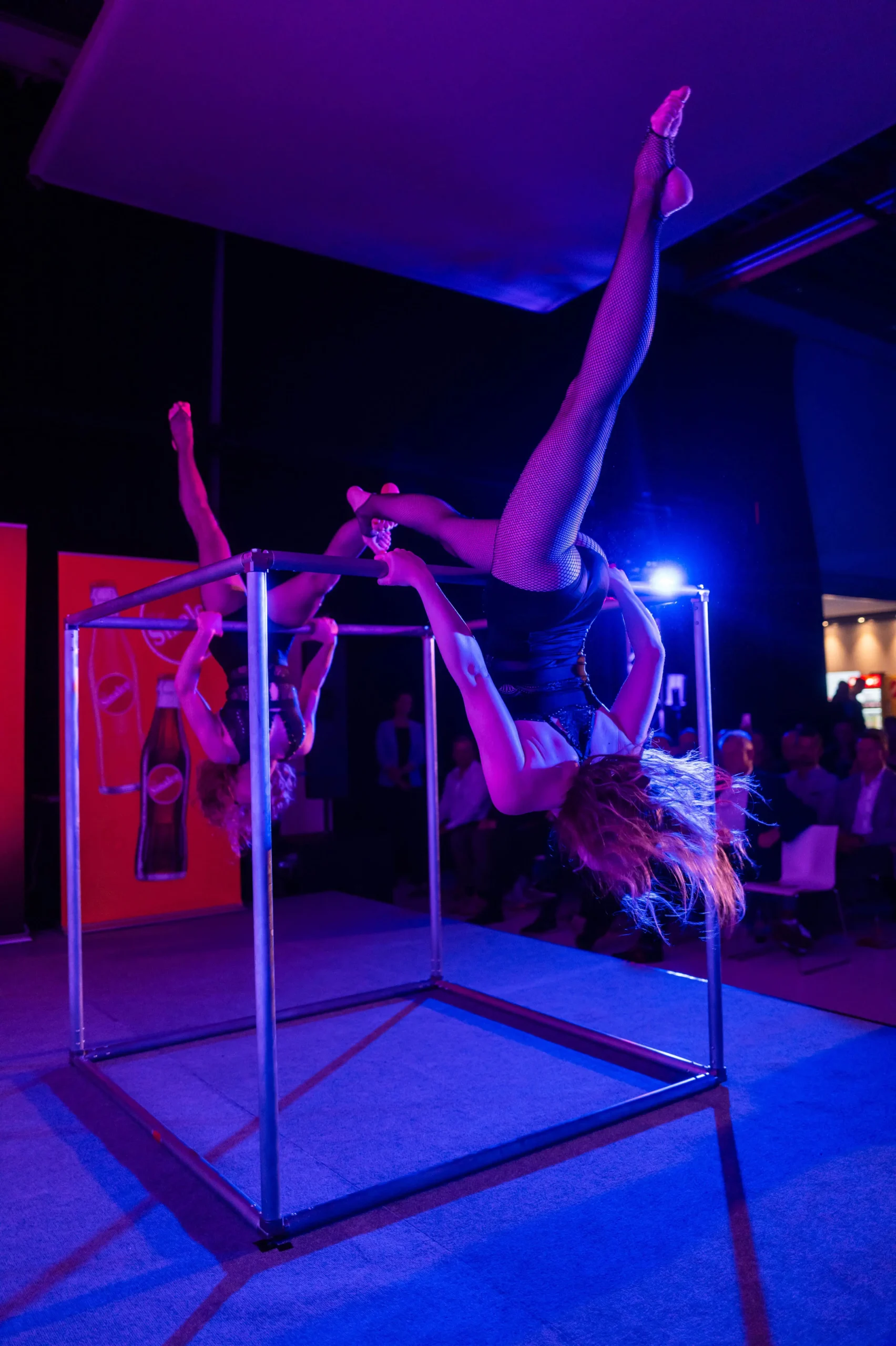 Zwei Akrobaten zeigen eine faszinierende Duo Acrobatic Cube Show, bei der sie mit ausgestreckten Beinen kopfüber auf einem Metallwürfel balancieren, der von blauer und violetter Bühnenbeleuchtung beleuchtet wird.