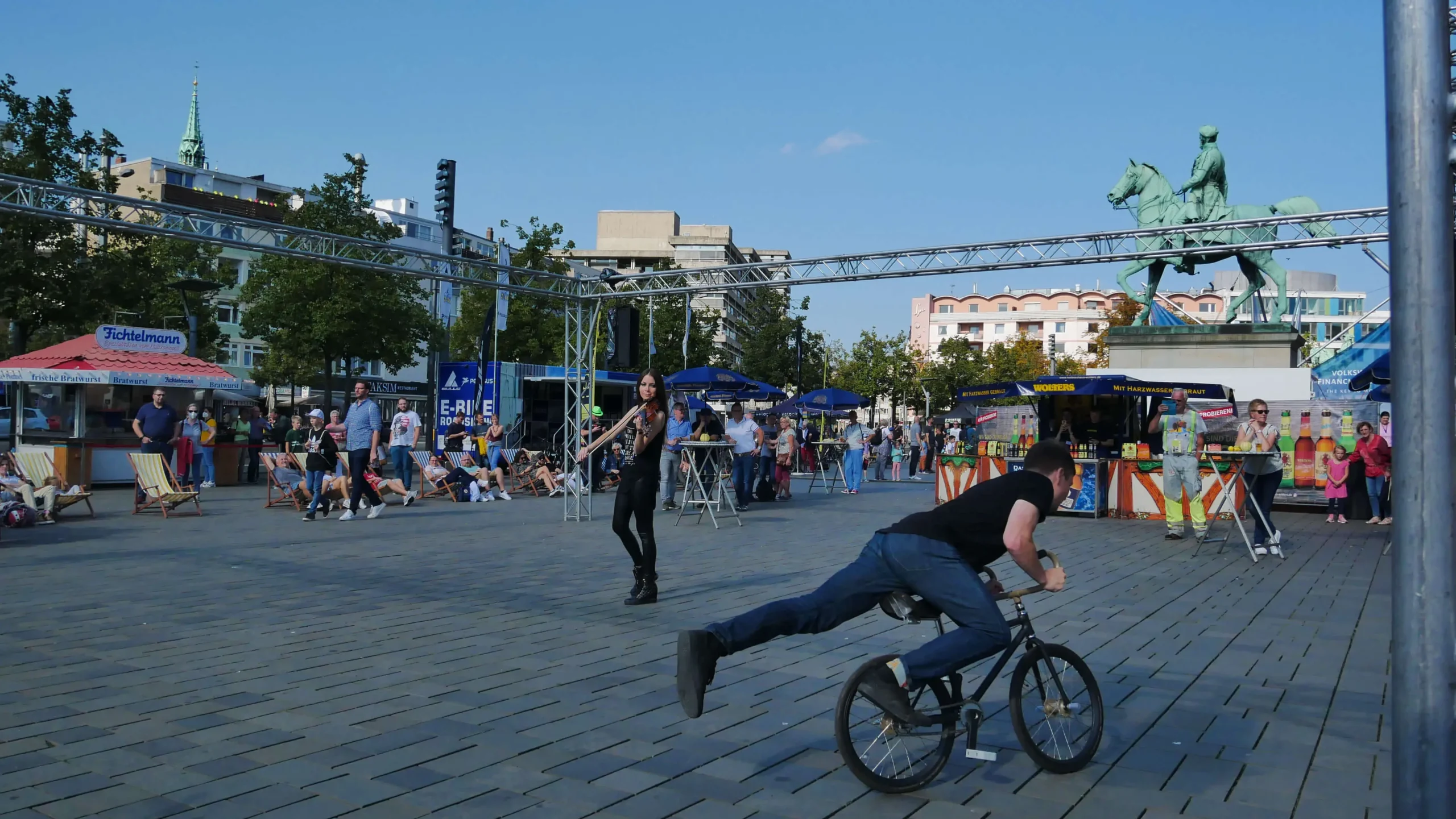 Ein Mann führt auf einem belebten städtischen Platz ein Kunststück auf einem BMX-Rad vor. Es entsteht das Gefühl einer improvisierten Violine & Fahrrad Show, während die Menschen zwischen Bäumen, Kiosken und einer großen Statue unter dem klaren blauen Himmel sitzen, spazieren gehen und zusehen.