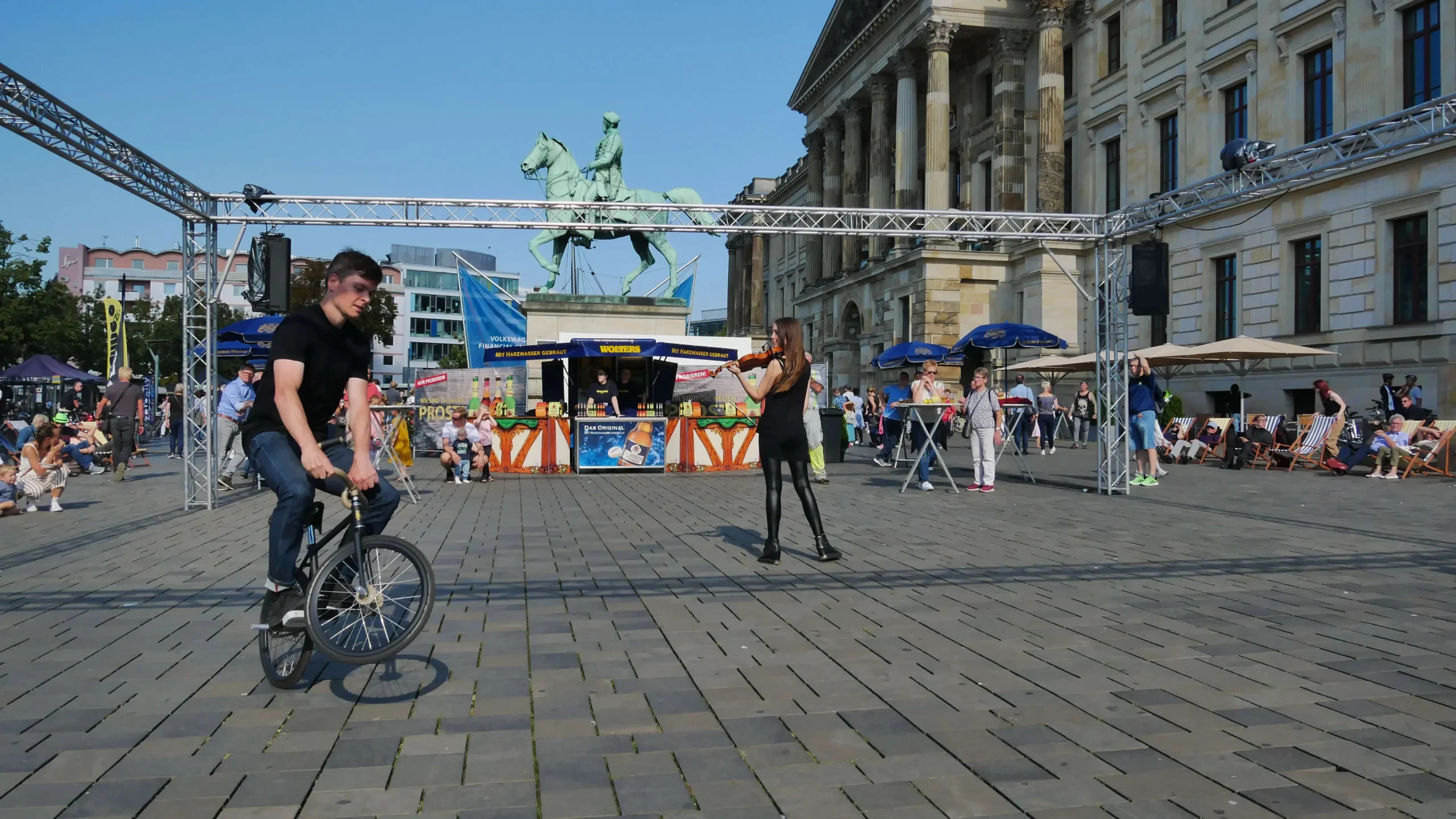 Ein Mann fährt mit dem Fahrrad auf einem Stadtplatz, während eine Frau mit einem Mikrofon die Violine & Fahrrad Show moderiert. Menschen sitzen unter Sonnenschirmen, und im Hintergrund sind ein großes Reiterstandbild und historische Gebäude zu sehen.