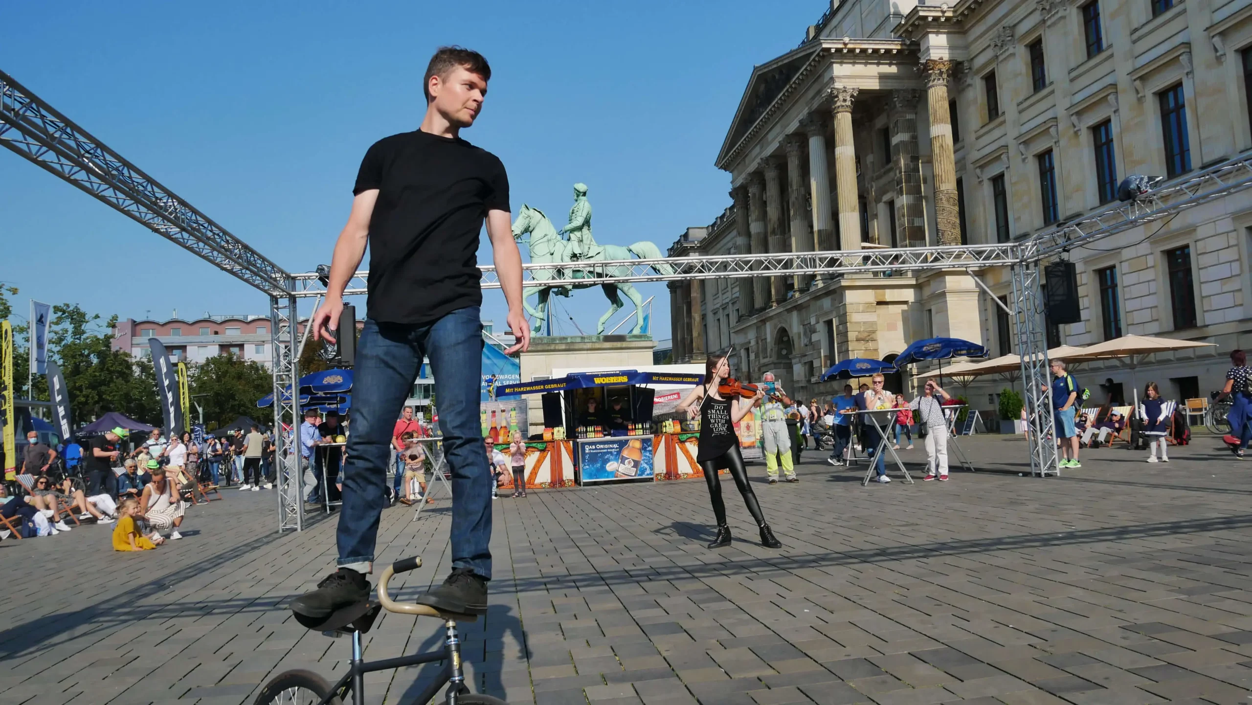 Ein junger Mann balanciert auf dem Lenker eines BMX-Rads auf einem belebten Stadtplatz während einer Violine & Fahrrad-Show, mit Spaziergängern und einer Statue eines Pferdes und eines Reiters im Hintergrund an einem sonnigen Tag.