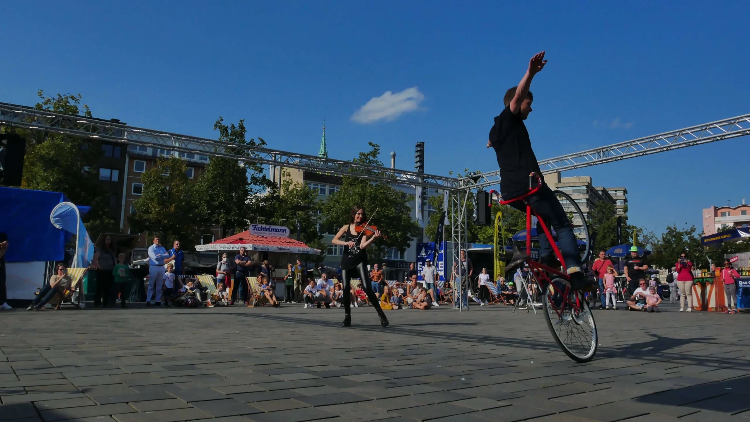 Ein Künstler balanciert auf einem Rad eines Fahrrads auf einem belebten Platz während der Violine & Fahrrad Show, während im Hintergrund Zuschauer unter einem klaren blauen Himmel sitzen und stehen. Ein anderer Artist steht in der Nähe.