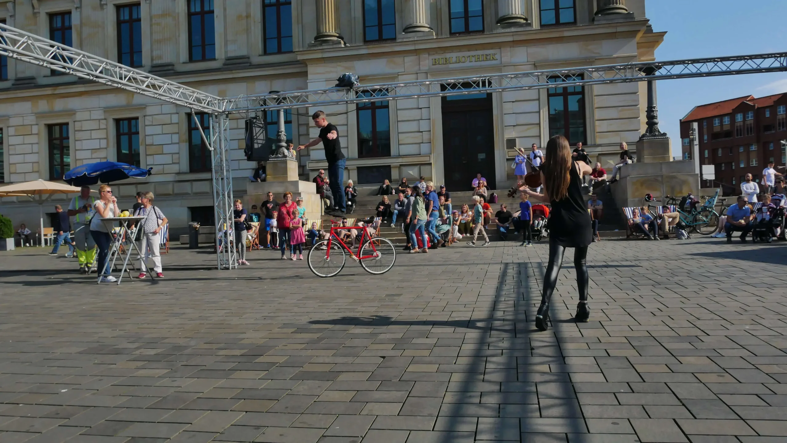 Ein Straßenkünstler der Violine & Fahrrad Show balanciert auf einem Drahtseil über einem roten Fahrrad, während eine Menschenmenge vor einem großen Gebäude zusieht. Eine schwarz gekleidete Frau steht der Künstlerin gegenüber, während im Hintergrund Menschen sitzen und stehen.