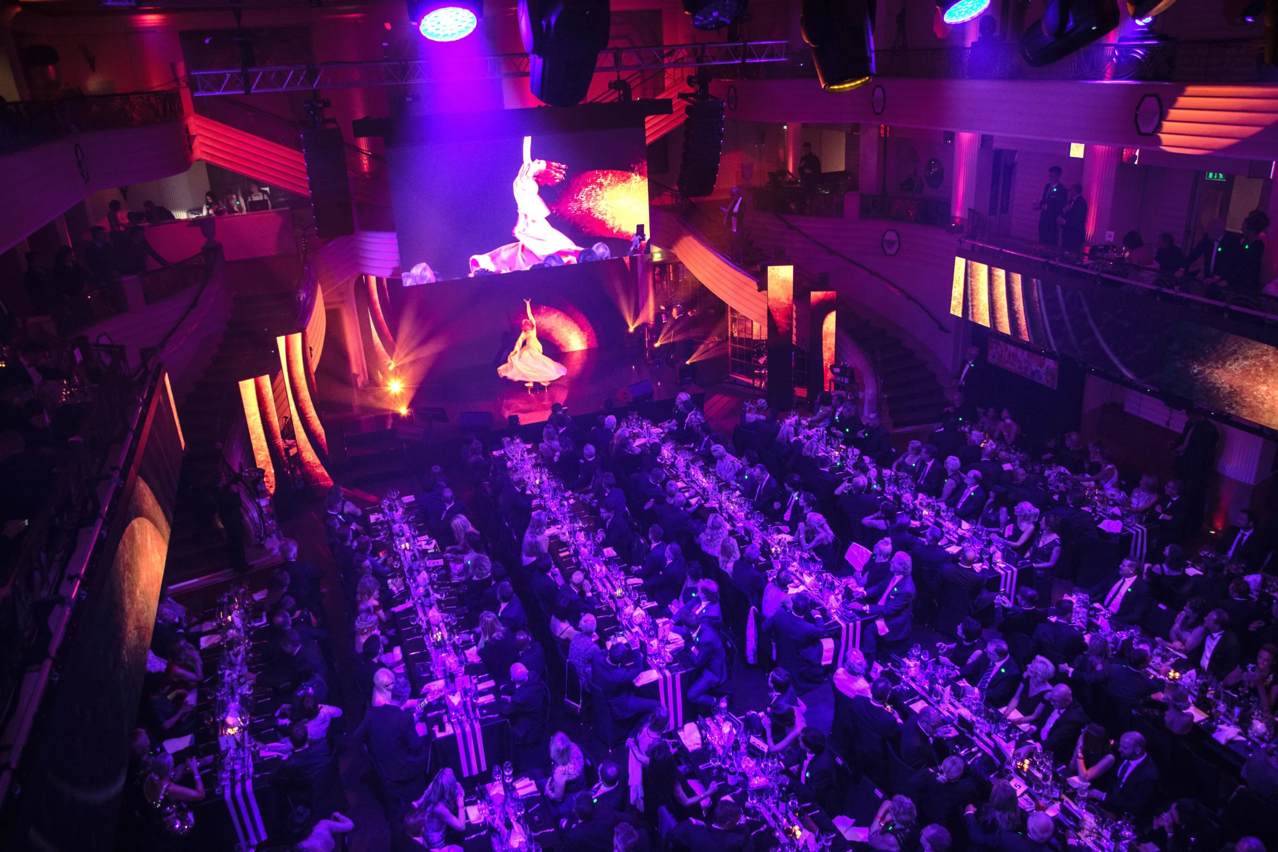 Ein großer, eleganter Bankettsaal für eine Firmenfeier, mit langen Tischen voller Gäste, dramatischer violetter und orangefarbener Beleuchtung und einer Bühne, auf der eine Darstellerin in einem weißen Kleid unter Scheinwerfern tanzt, während ihr Bild auf einer großen Leinwand darüber angezeigt wird.
