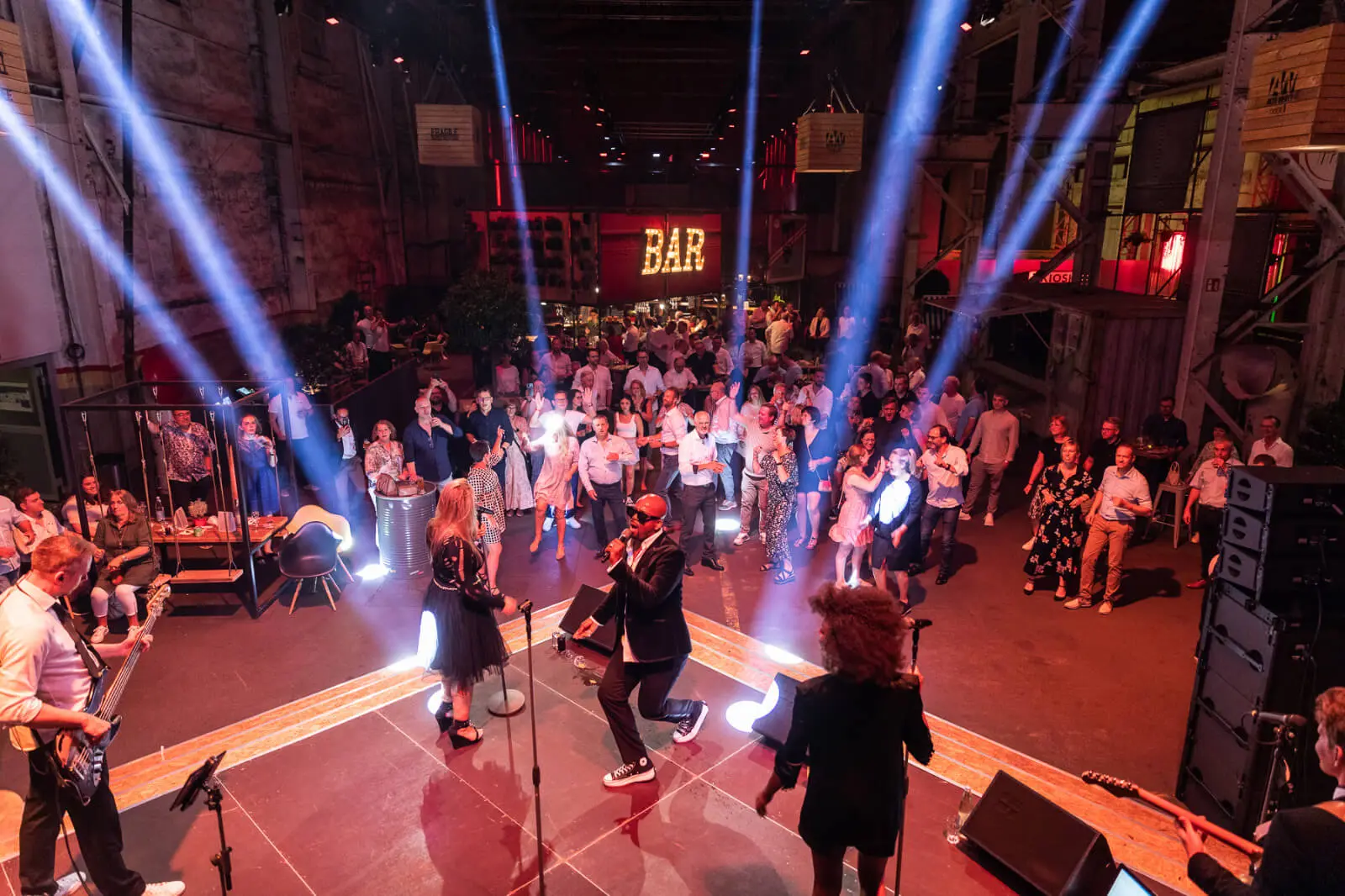 Eine Live-Band heizt einer Firmenfeier auf der Bühne vor einer großen Menschenmenge in einer Halle ein, während die BAR im Hintergrund beleuchtet ist. Helle Bühnenlichter erhellen die Gäste, die tanzen und sich unter die Gäste mischen, und sorgen für eine unvergessliche Firmenfeier.