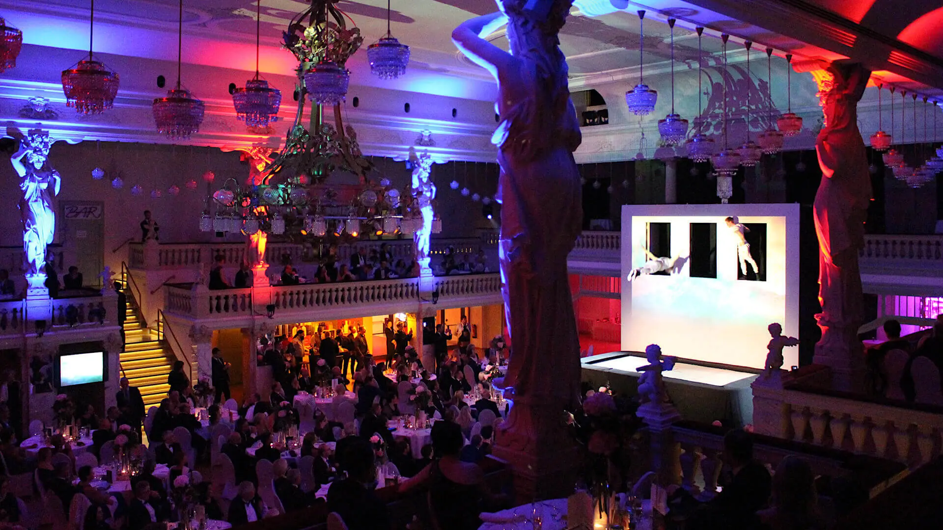 Ein großer, eleganter, mit bunten Lichtern beleuchteter Ballsaal, gefüllt mit Gästen, die an runden Tischen sitzen und eine festliche Firmenfeier veranstalten. Statuen umgeben den Raum, und im Hintergrund ist eine Bühne mit einer hellen Aufführung zu sehen.