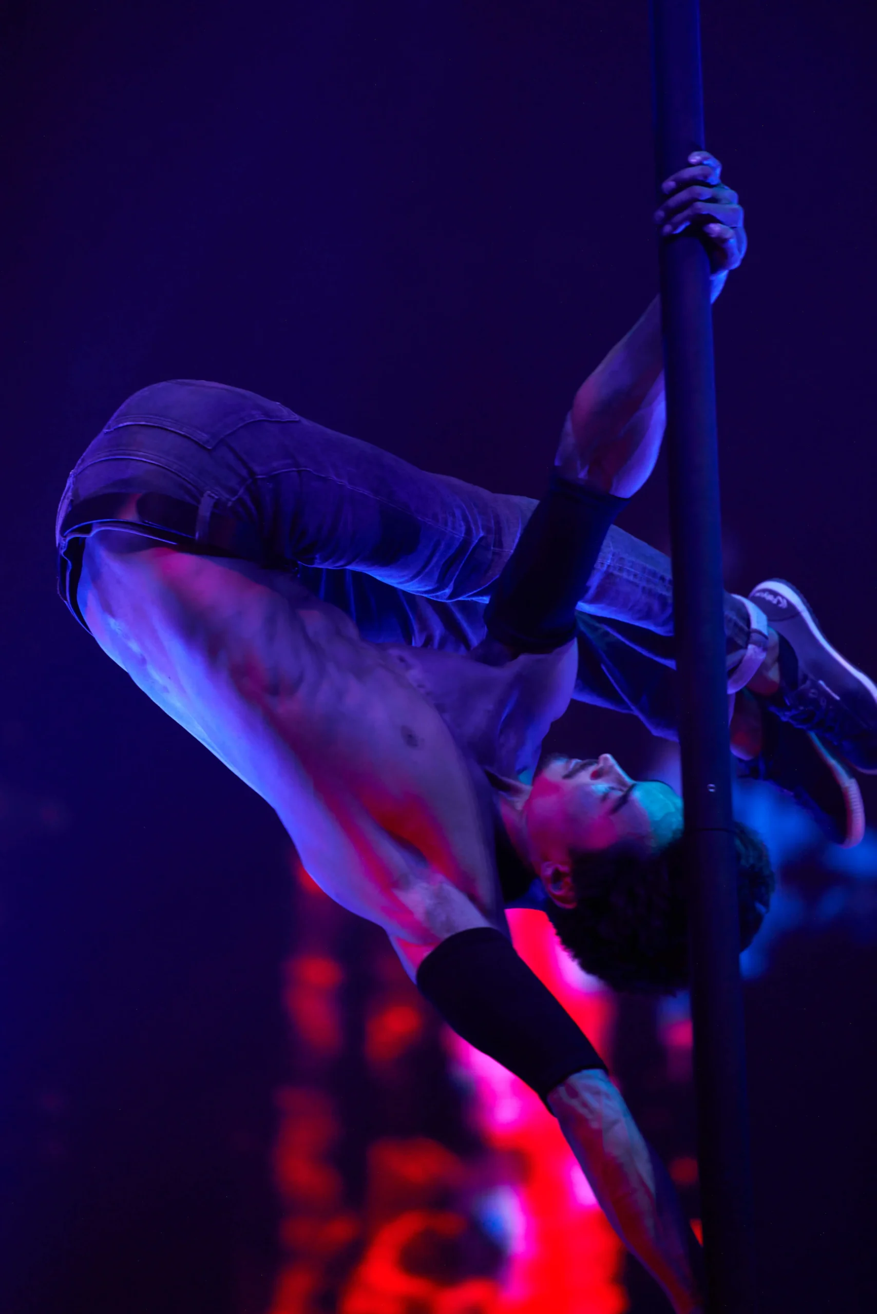 Eine hemdsärmelige Person in Jeans und Turnschuhen vollführt eine akrobatische Bewegung an einer vertikalen Stange, wobei sie sich mit Armen und Beinen festhält, während sie kopfüber in einer dramatischen blauen und roten Bühnenbeleuchtung während einer Flying Pole Show schwebt.