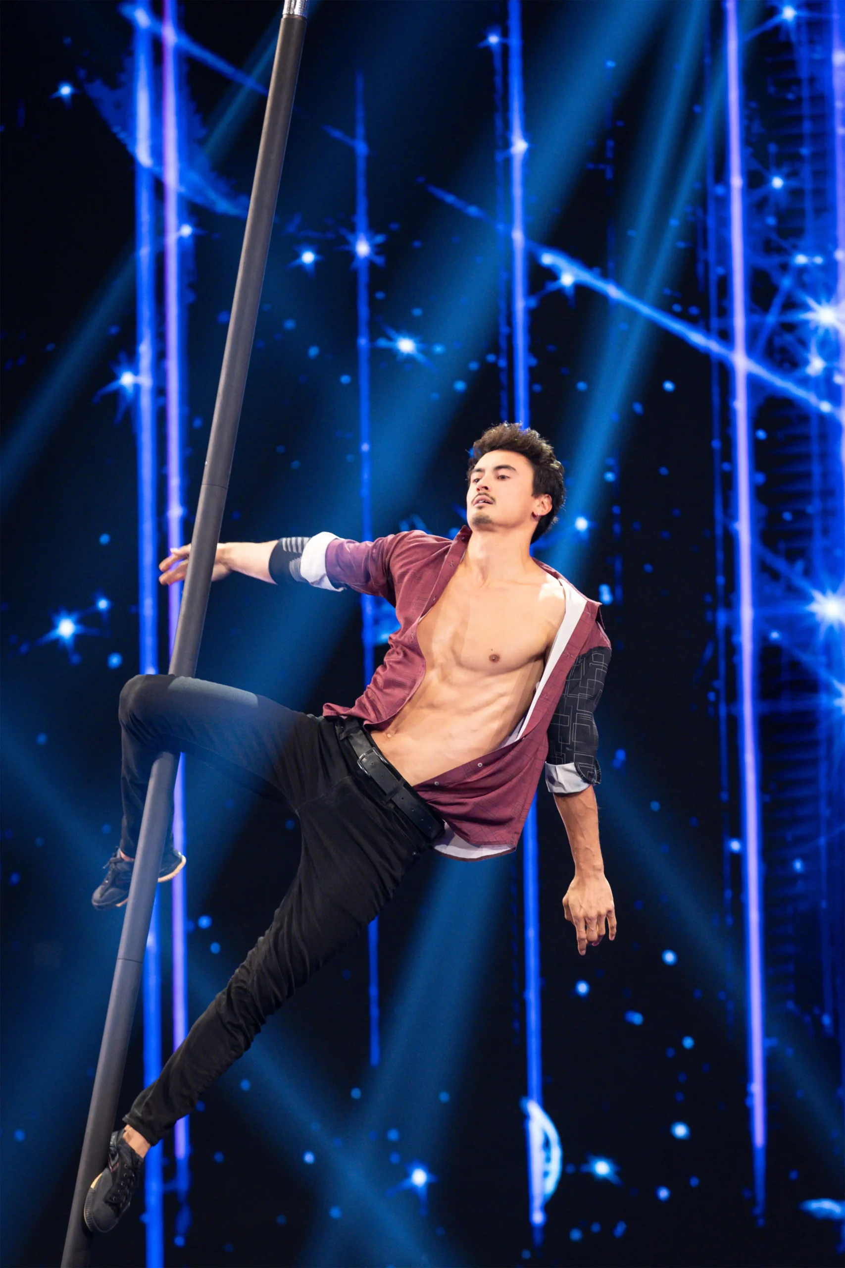 Ein männlicher Stangenakrobat in schwarzer Hose und offenem kastanienbraunem Hemd führt auf der Bühne eine dramatische Flying Pole Show vor, bei der er sich mit einem Arm und einem Bein an einer vertikalen Stange festhält, vor einer Kulisse aus blauen Lichtern und sternähnlichen Effekten.