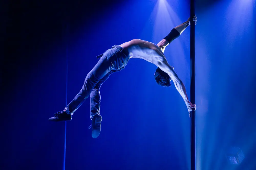 Ein hemdsärmeliger Darsteller führt während einer Flying Pole Show eine dramatische Pose in der Luft aus, indem er die vertikale Stange mit einem Arm umgreift und seinen Körper unter blauer Bühnenbeleuchtung horizontal ausstreckt.