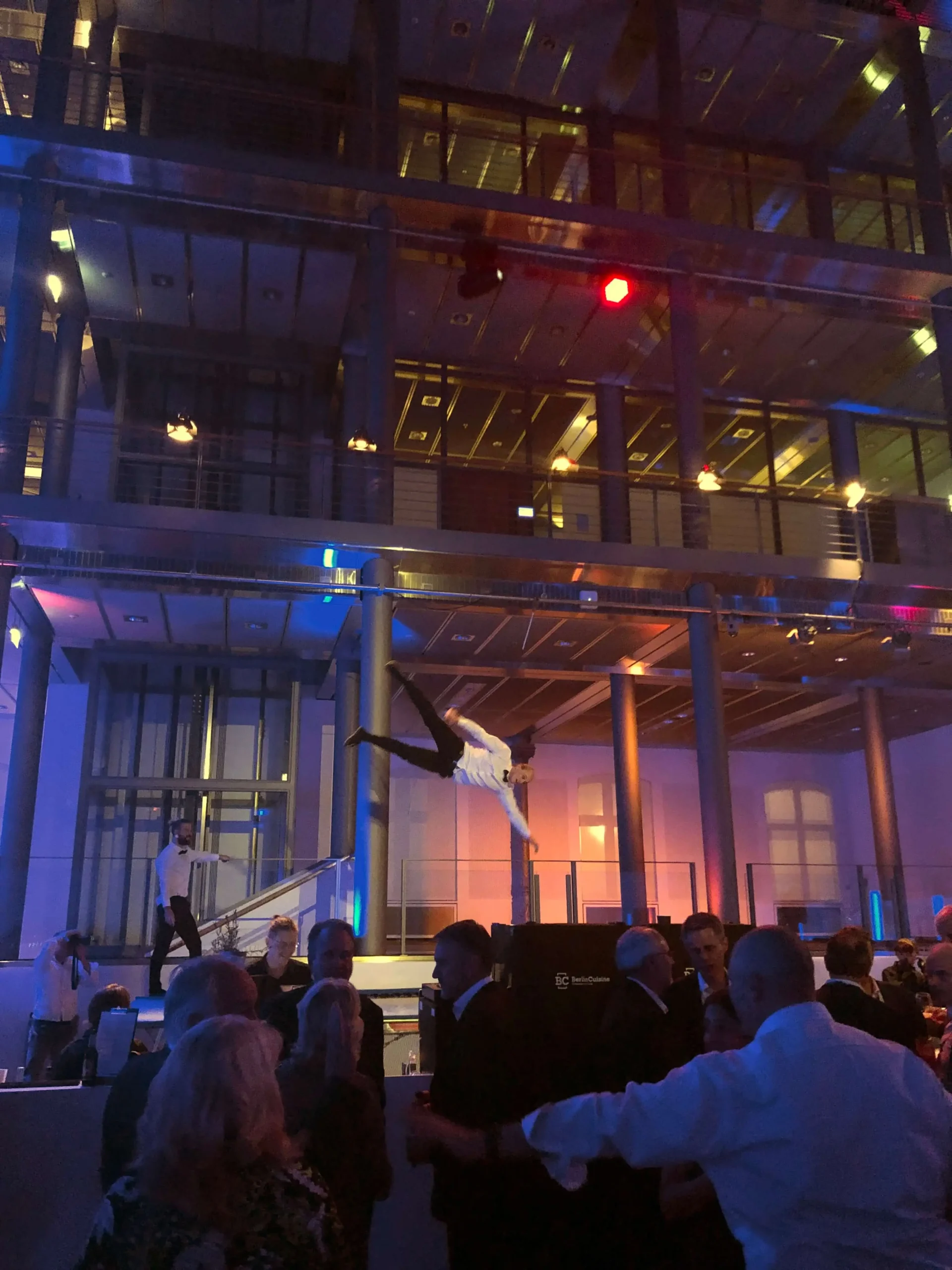 Ein Artist springt in der Luft über einer Menschenmenge bei einer Indoor-Gala, während im Hintergrund ein modernes Glasgebäude von bunten Lichtern beleuchtet wird. Die Menschen unten beobachten die Akrobatikshow.