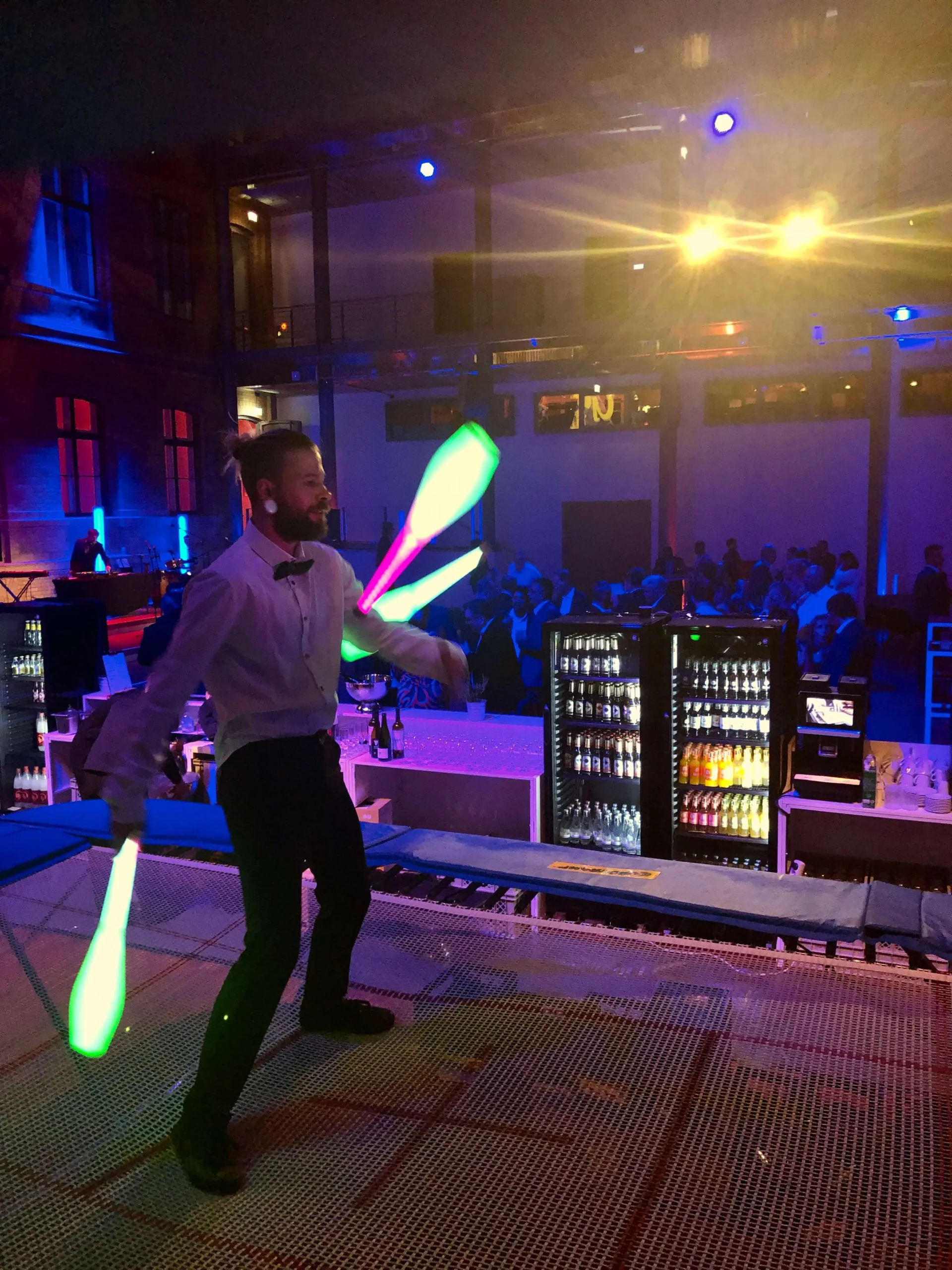 Auf einer lebhaften Indoor-Gala jongliert ein Mann in weißem Hemd und Fliege mit leuchtenden Keulen auf einem Trampolin, während die Menge im Hintergrund zusieht.