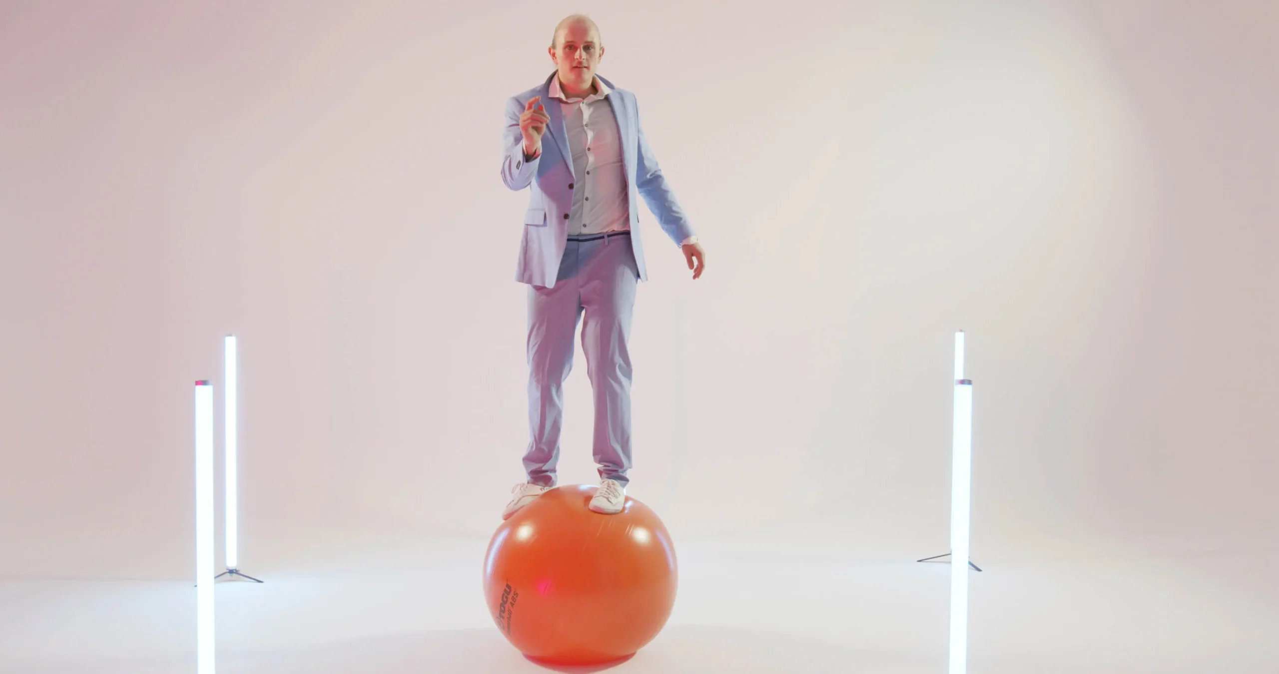 Ein Mann in einem hellblauen Anzug balanciert auf einem orangefarbenen Gymnastikball in einem minimalen, hell beleuchteten Raum mit zwei vertikalen Lichtröhren auf beiden Seiten, die eine eindrucksvolle Gym Ball Show-Atmosphäre schaffen.