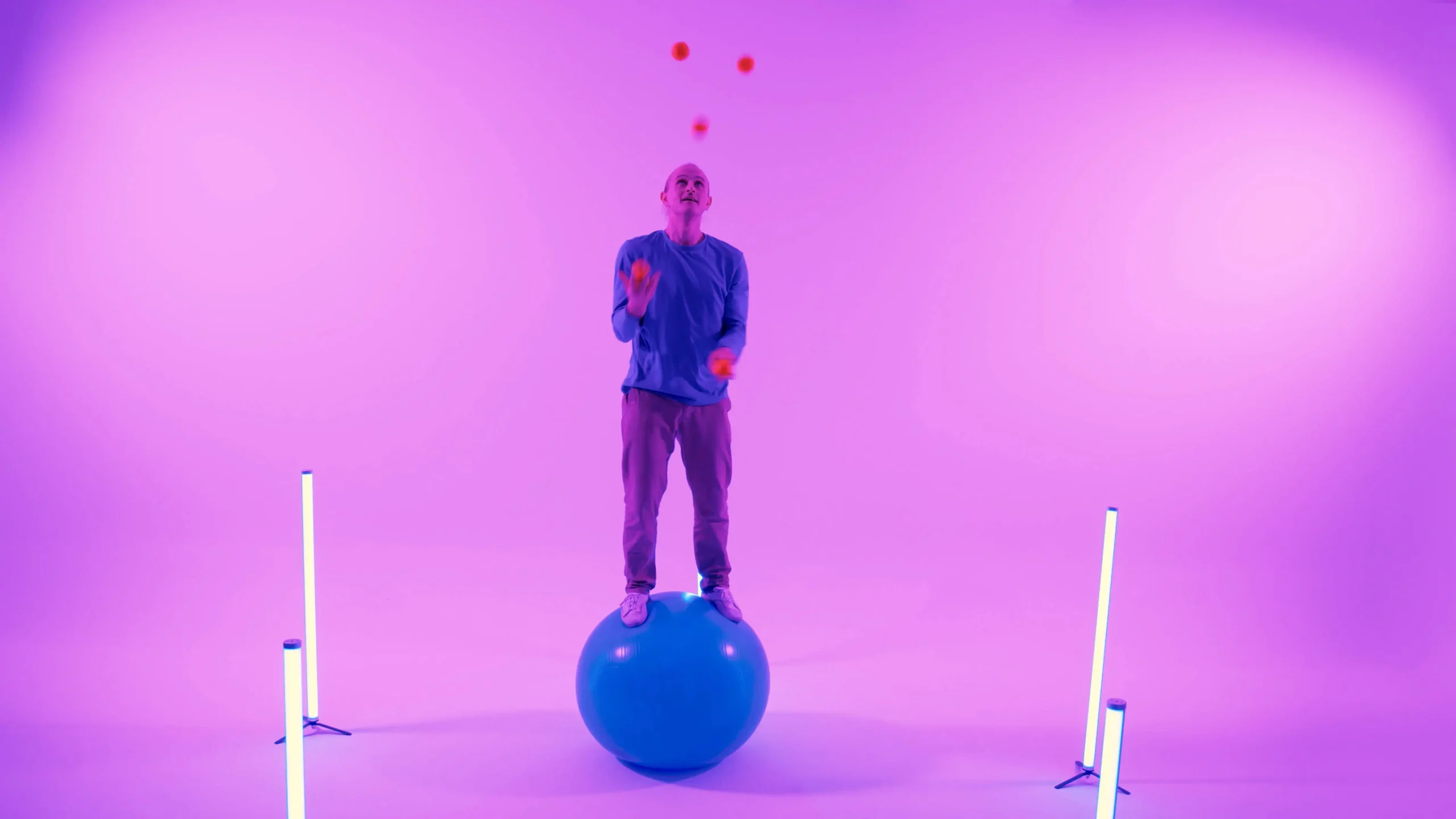 Eine Person führt eine Gymnastikball-Show vor, bei der sie auf einem großen blauen Ball steht und mit orangefarbenen Bällen in einem mit lila und rosa Lichtern beleuchteten Raum jongliert, mit zwei vertikalen Lichtbalken auf beiden Seiten.