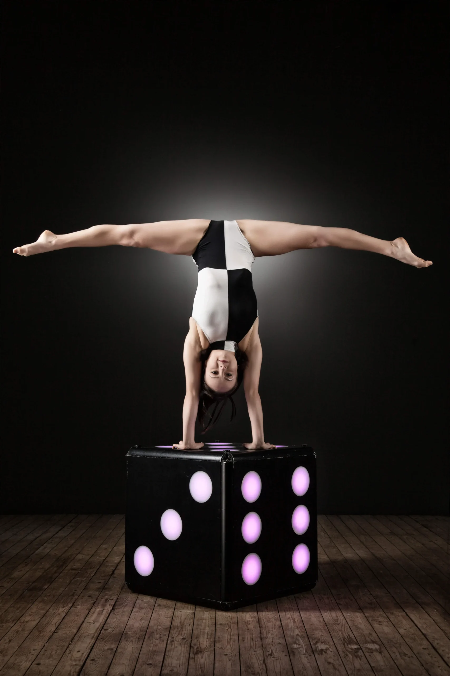 Eine Frau in einem schwarz-weißen Trikot zeigt atemberaubende Handstandakrobatik, indem sie einen breitbeinigen Handstand auf einem großen, beleuchteten Würfel auf einem Holzboden vor einem dunklen Hintergrund vorführt.