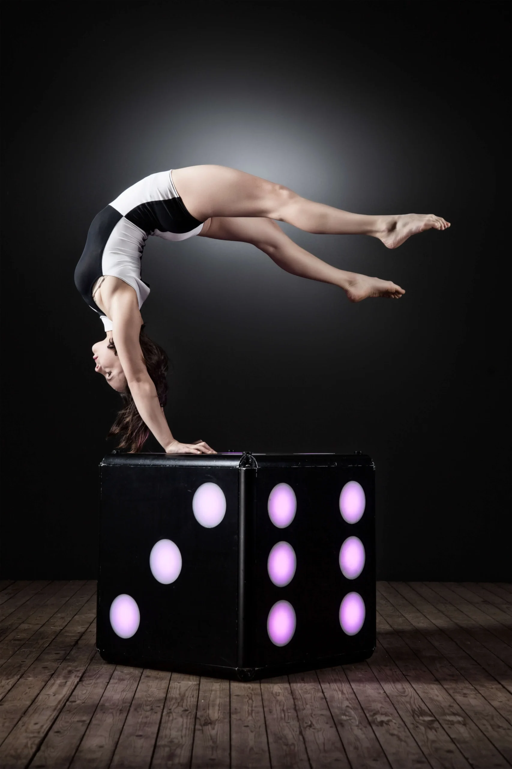 Eine Frau in schwarz-weißem Outfit zeigt Handstandakrobatik mit gewölbtem Rücken und balanciert auf einem großen beleuchteten Würfel vor einem dunklen Hintergrund auf einem Holzboden.