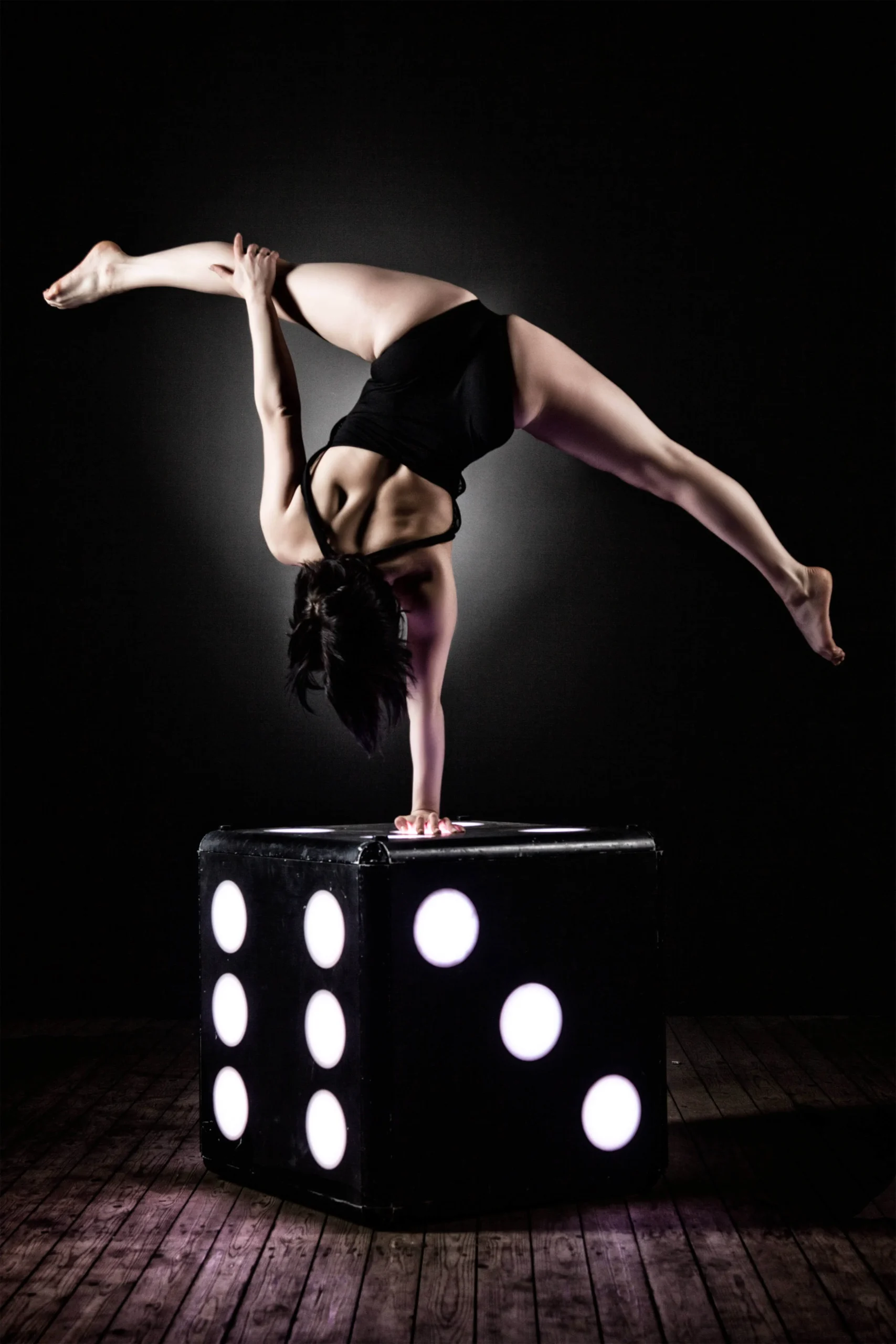 Eine Akrobatin in einem schwarzen Trikot zeigt Handstandakrobatik, indem sie einen einhändigen Handstand auf einem großen schwarzen Würfel mit weißen Punkten vollführt und dabei anmutig mit einem Bein nach oben und dem anderen zur Seite vor einem dunklen Hintergrund balanciert.