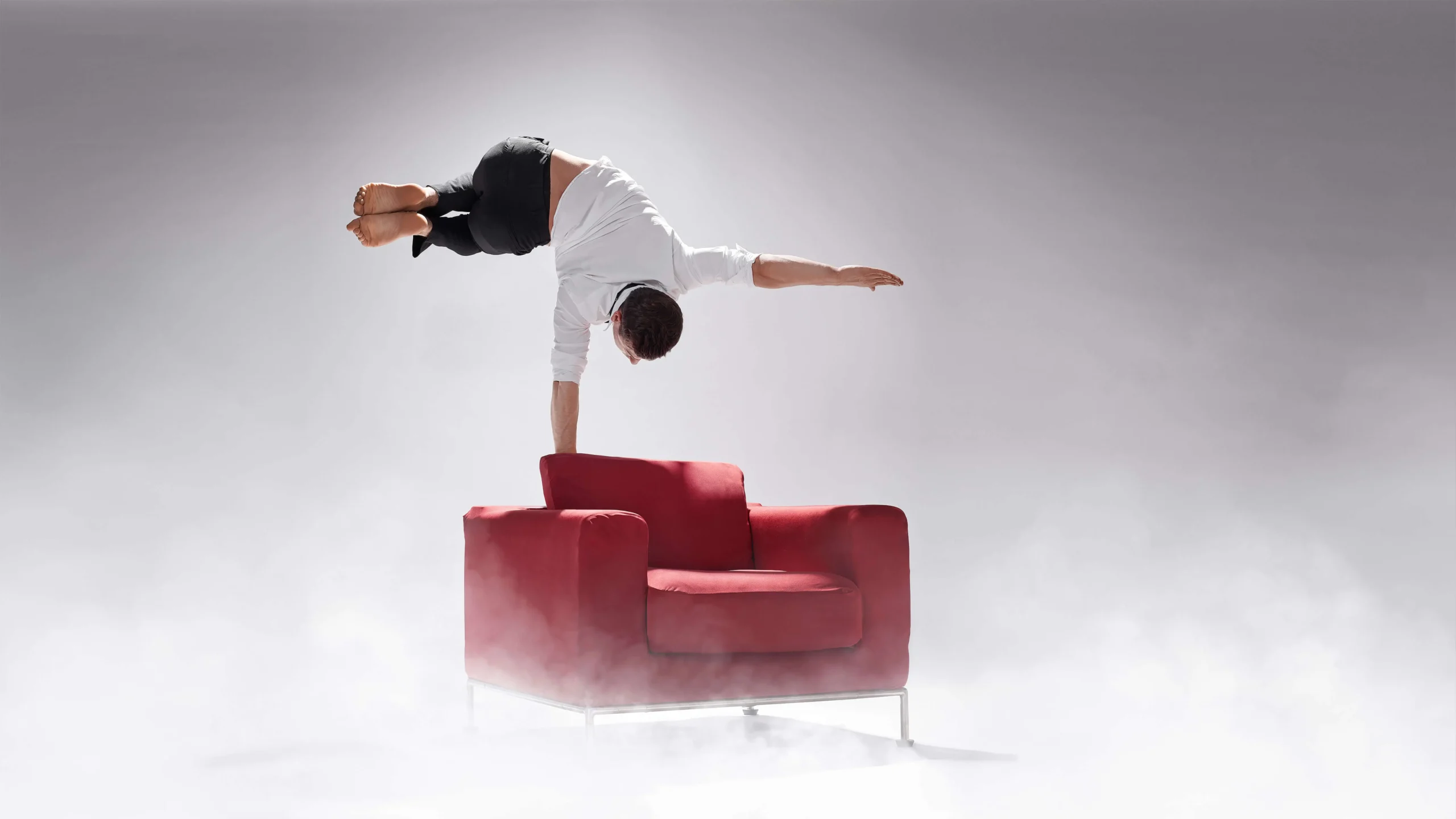 Eine mit weißem Hemd und dunkler Hose bekleidete Person vollführt eine beeindruckende Handstandakrobatik, indem sie kopfüber auf einer Hand auf der Armlehne eines roten Sessels balanciert, umgeben von Nebel oder Dunst vor einem hellgrauen Hintergrund.