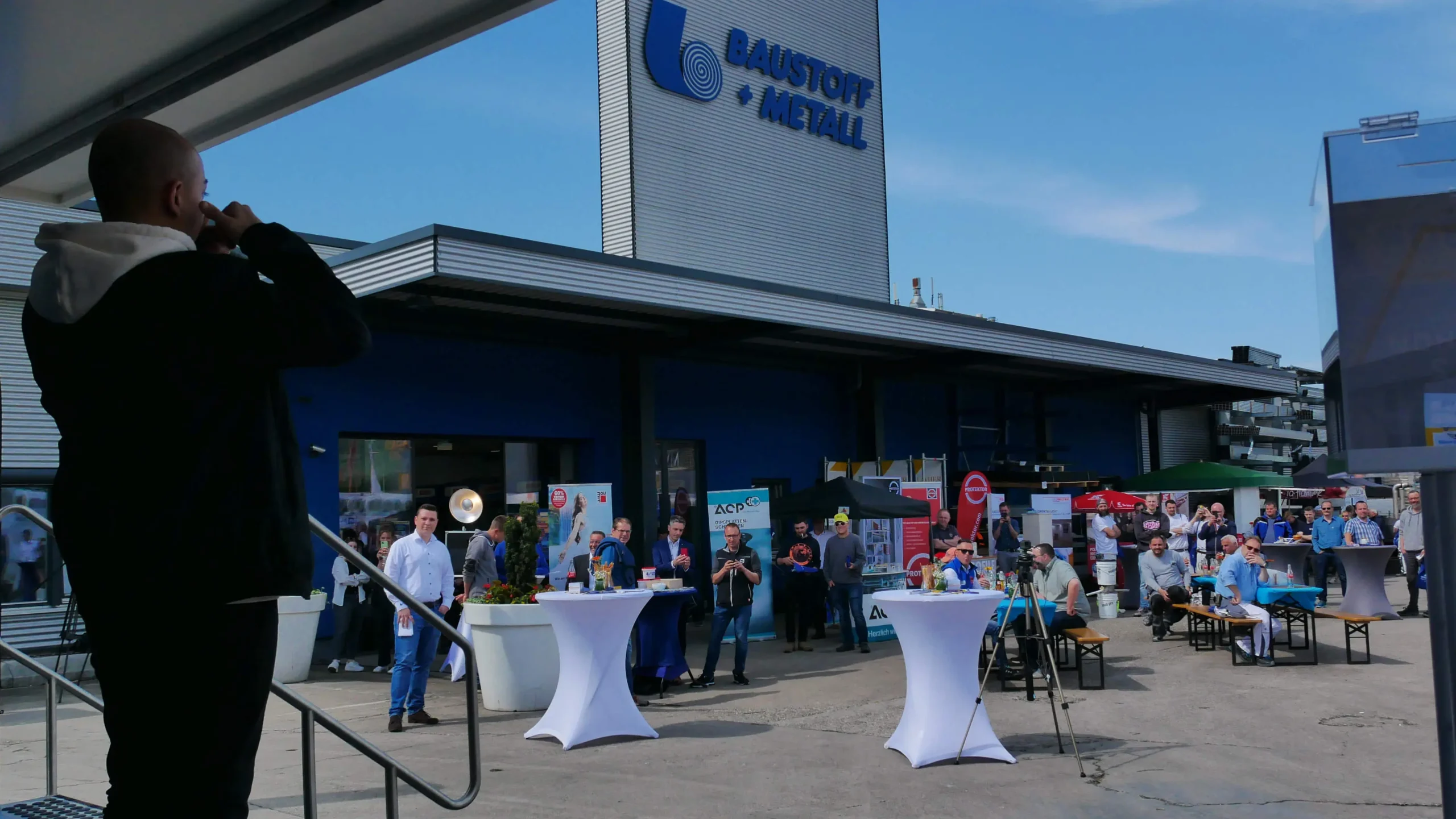 Eine Person spricht in ein Mikrofon auf einer Bühne vor einem Gebäude mit der Aufschrift BAUSTOFF METALL während eines Messeauftritts im Freien, wo sich Menschen um Cocktailtische und Informationsstände scharen.