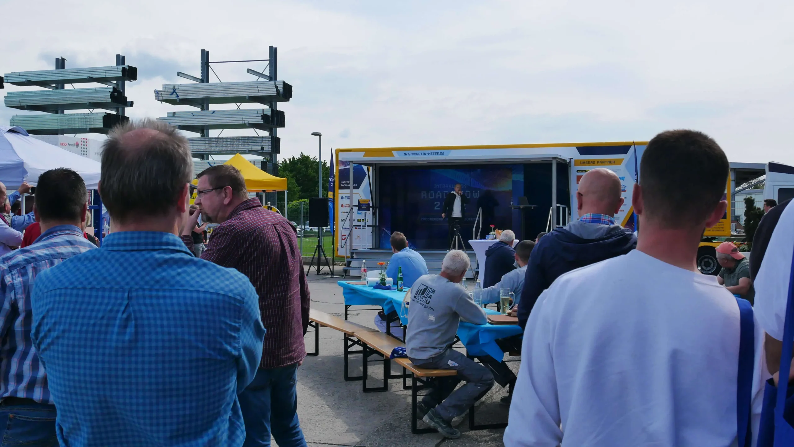 Eine Gruppe von Menschen versammelt sich im Freien an Picknicktischen mit blauen Tischtüchern und nimmt an einem Messeauftritt teil, bei dem ein Redner von einer in einem gelben Anhänger aufgebauten Bühne aus präsentiert. Im Hintergrund sind Baumaterialien gestapelt.