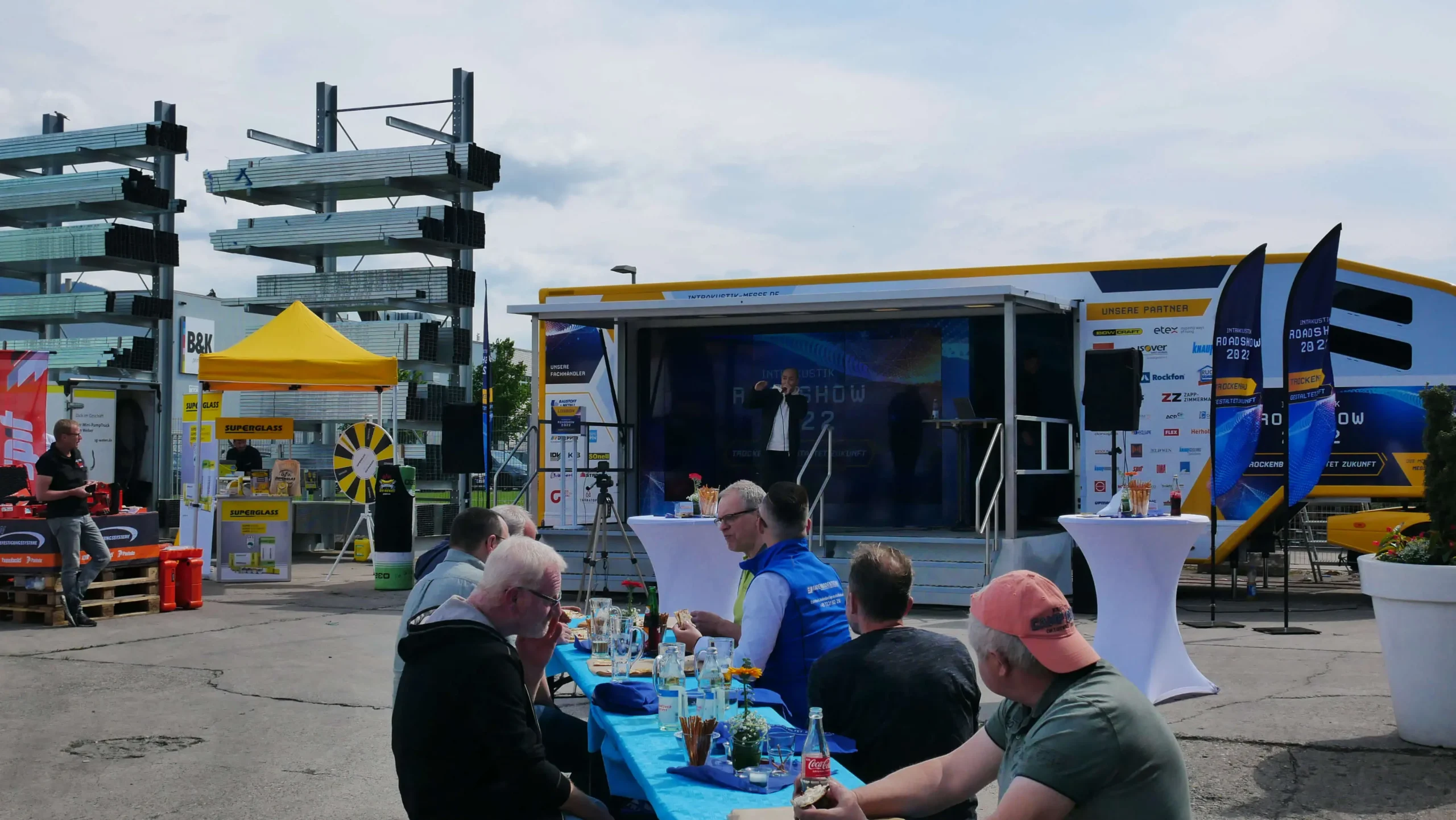 Die Menschen sitzen an einem langen Tisch im Freien mit Speisen und Getränken und genießen ein Bühnenprogramm vor einem gelb-weißen Lastwagen - ein lebendiger Messeauftritt, eingerahmt von Werbebannern und Industriematerialien im Hintergrund.