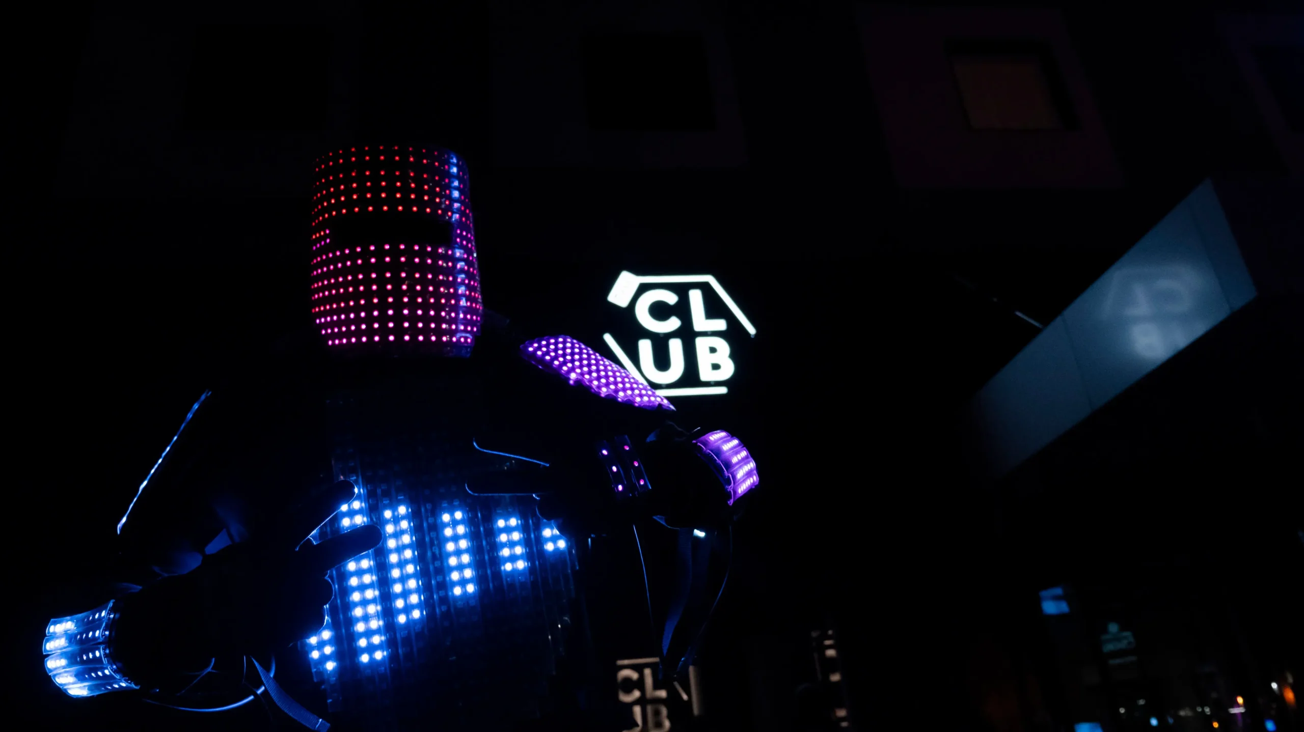 Eine Person in einem futuristischen LED-Roboter-Kostüm, das mit bunten Lichtern bedeckt ist, steht vor einem Gebäude mit einem leuchtenden CLUB-Schild und schafft eine lebendige und hochtechnologische Atmosphäre bei Nacht.