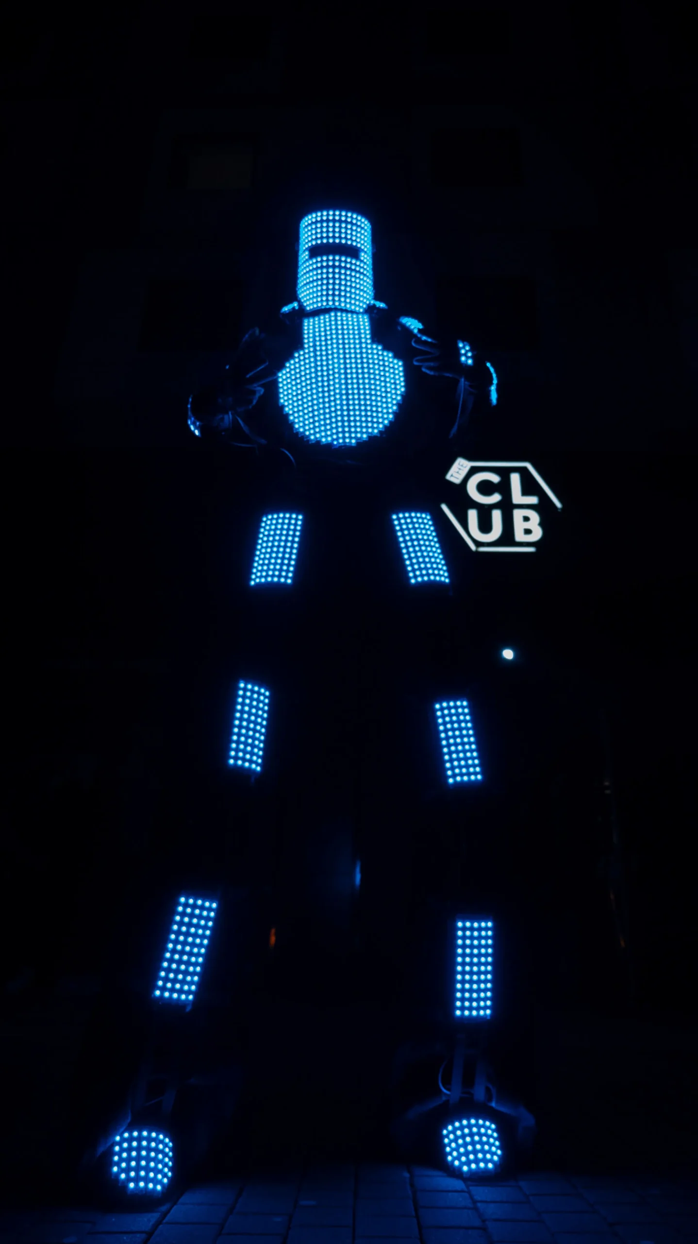 Ein LED-Roboter in einem futuristischen Anzug mit hellen blauen LED-Lichtern steht vor einem dunklen Hintergrund mit einem teilweise sichtbaren CLUB-Schild.