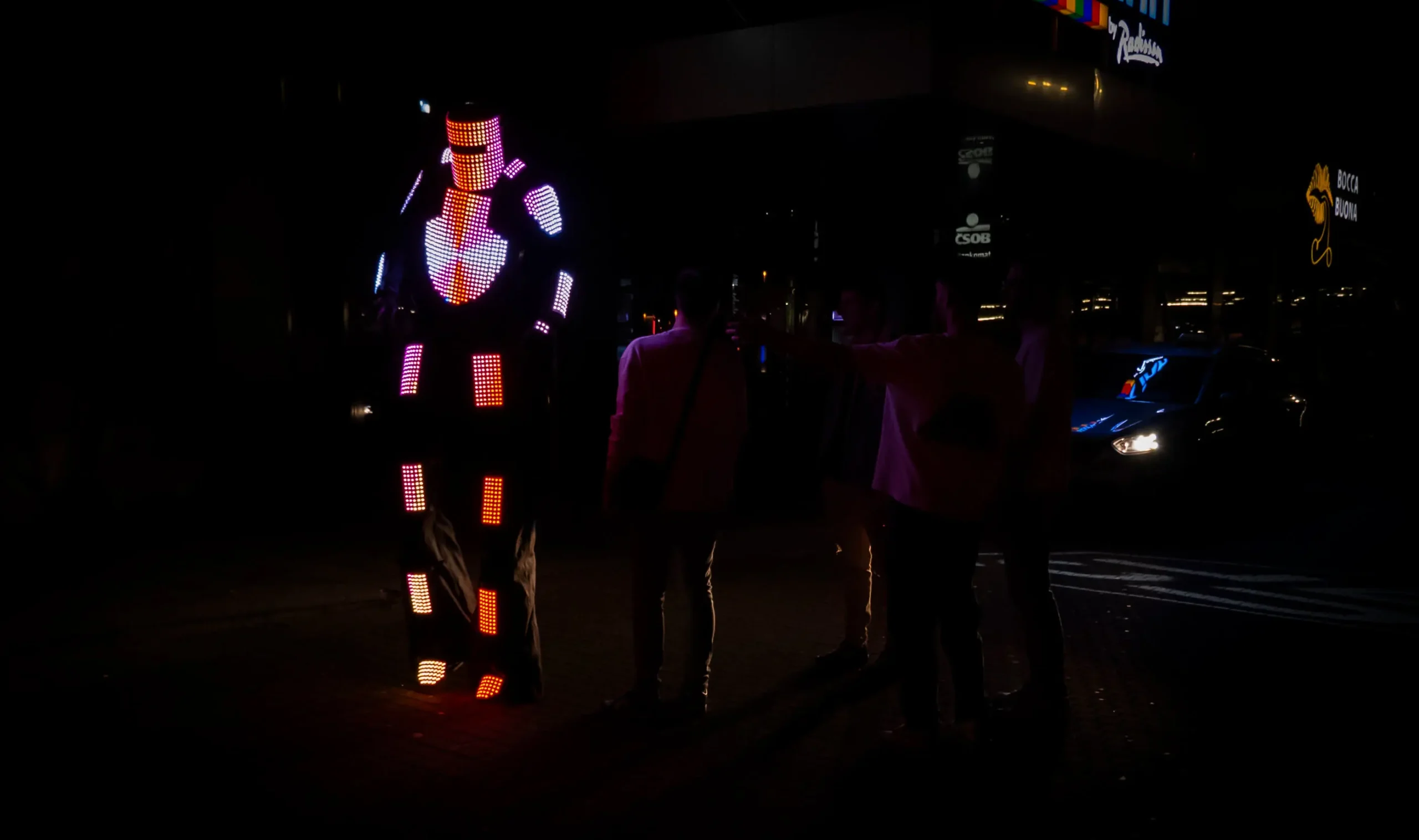 Eine Person in einem leuchtenden, mehrfarbigen LED-Roboteranzug steht in der Dunkelheit, während mehrere Personen im Schatten zusehen und Fotos machen. Im Hintergrund schimmern ein Auto und die Lichter der Stadt.
