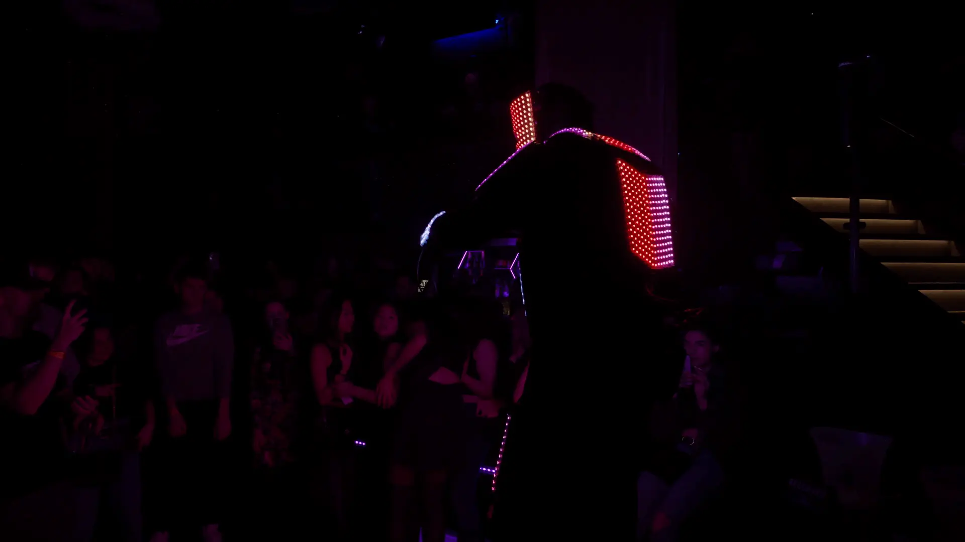 Ein als LED-Roboter verkleideter Darsteller steht in einem dunklen Raum, sein Kostüm leuchtet mit roten LED-Lichtern, die vertikale Linien auf seinem Rücken bilden. Das Publikum schaut zu, einige stehen, andere sitzen in der Nähe der beleuchteten Treppe auf der rechten Seite.