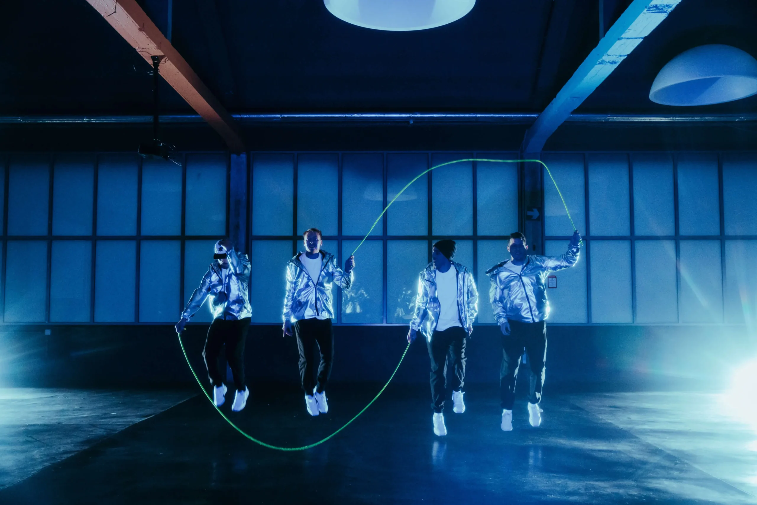 Vier Personen in glänzenden silbernen Jacken springen in einem dunklen Raum bei blauer Beleuchtung gemeinsam Seil, während sie ein großes grünes Springseil springen und drehen.