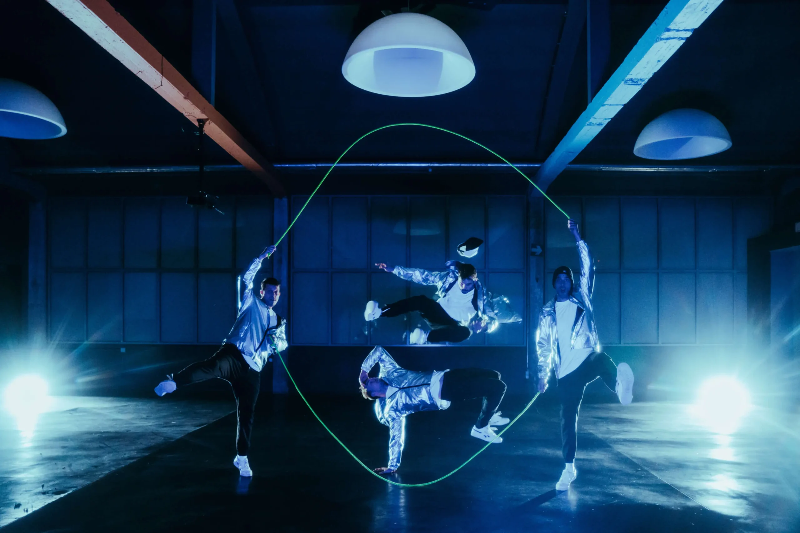 Vier Tänzerinnen und Tänzer in glänzenden silbernen Jacken führen in einem dunklen, blau beleuchteten Raum eine energiegeladene Seilsprungnummer auf, wobei zwei das Seil halten, während die anderen springen und in der Luft posieren. Helle Lichter erhellen den Hintergrund.