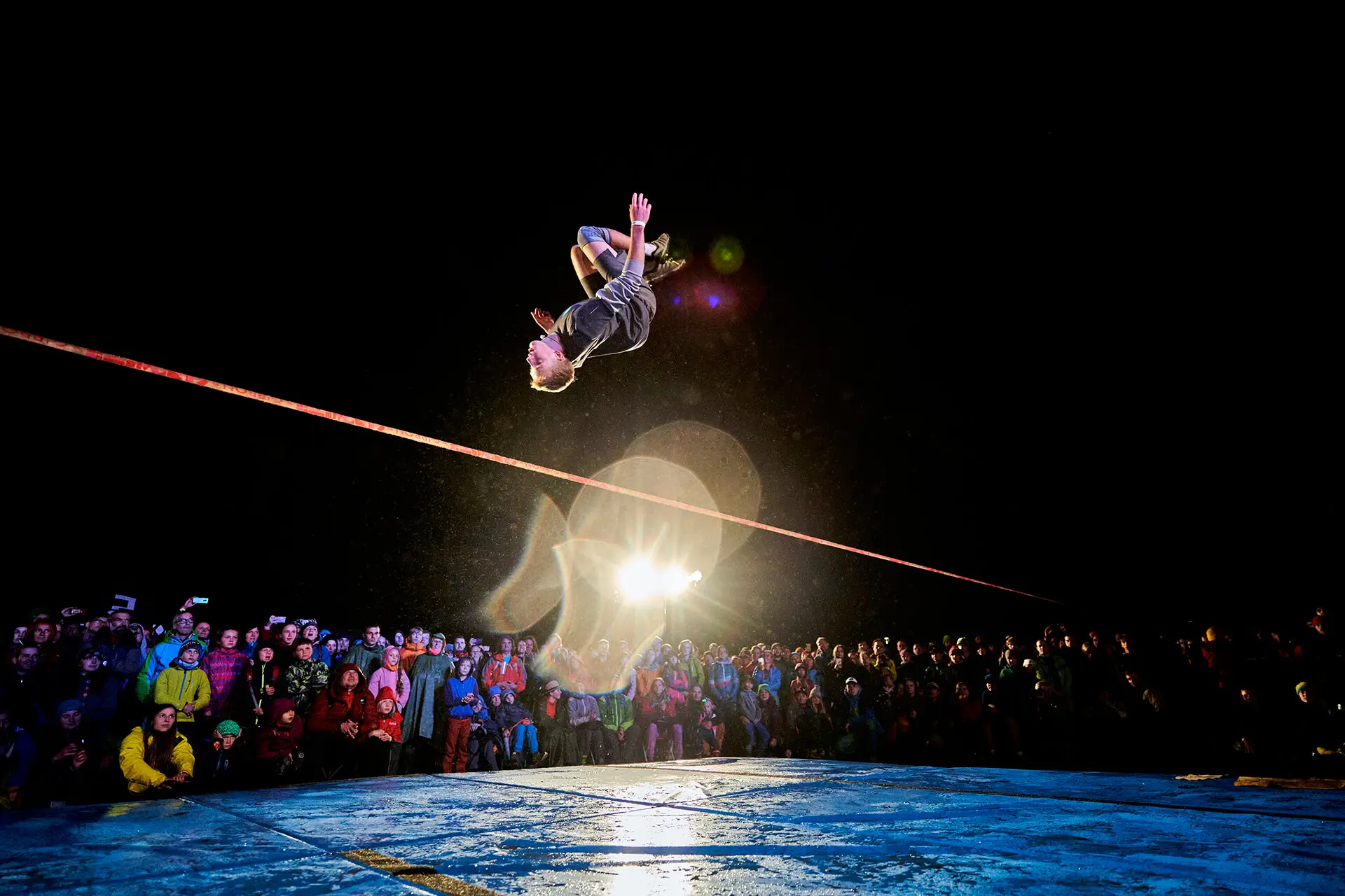 Eine Person vollführt einen waghalsigen Rückwärtssalto auf einer Slackline über einer großen Menschenmenge bei Nacht, während helle Lichter die Szene beleuchten und die Zuschauer von unten aufmerksam zusehen.