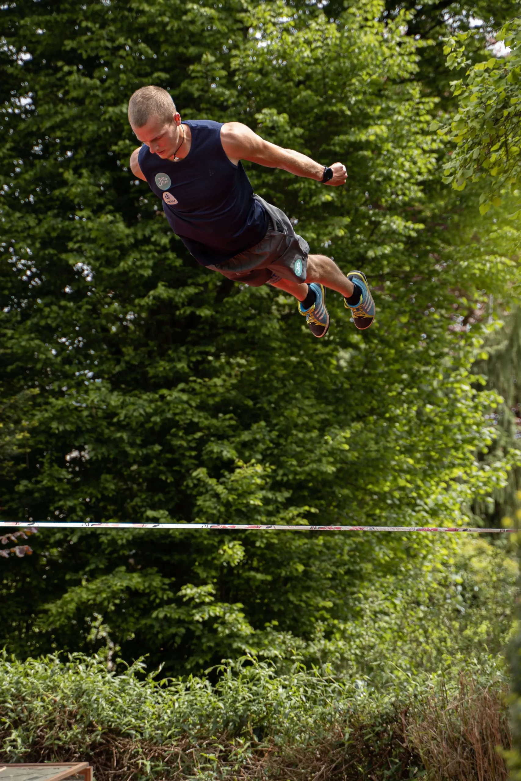 Ein Mann in sportlicher Kleidung wird in der Luft über einer Slackline, parallel zum Boden, vor dem Hintergrund dichter grüner Bäume gefilmt, um seinen gewagten Slackline-Trick oder -Sprung zu unterstreichen.