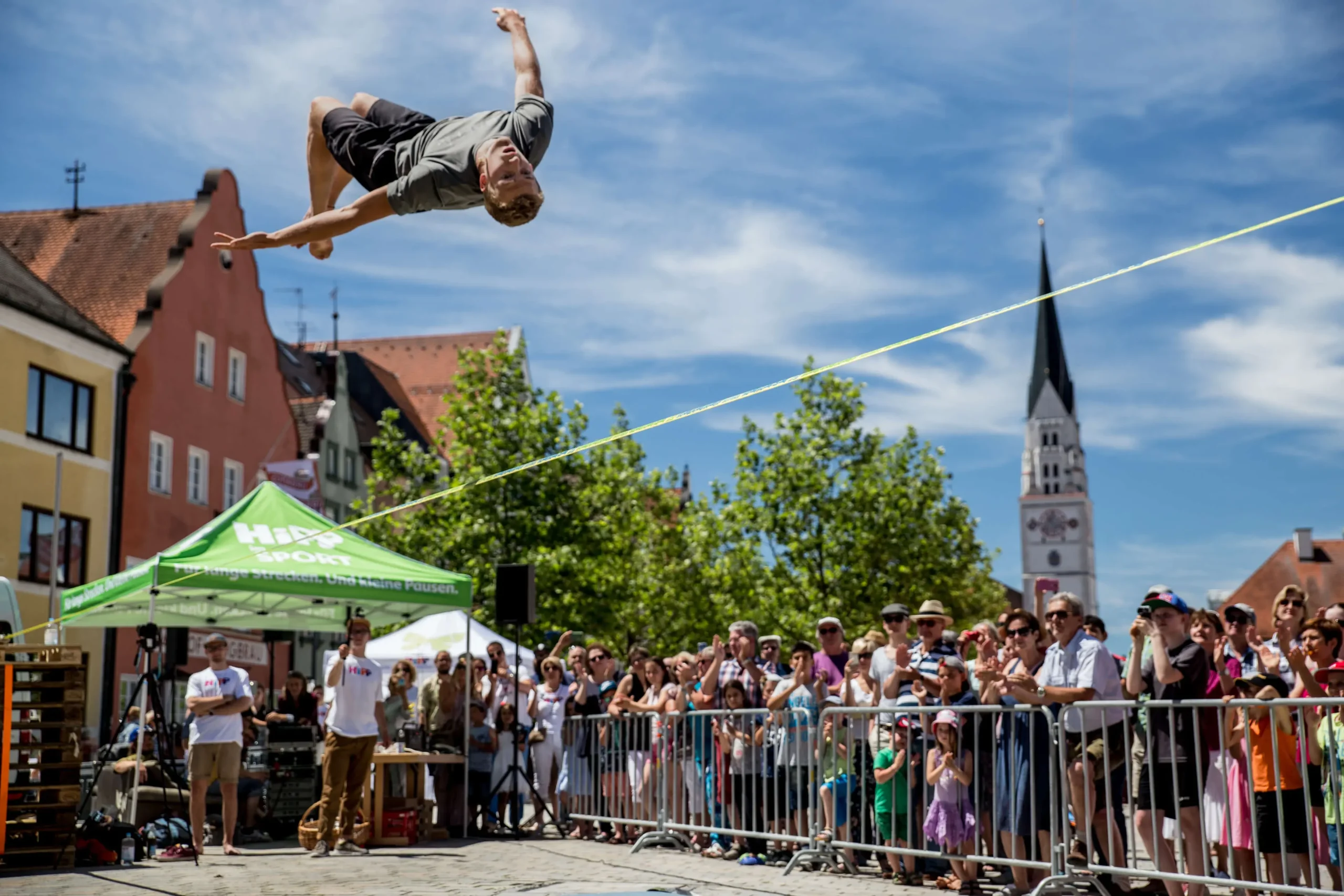 Ein Mann vollführt einen Rückwärtssalto auf einer Slackline bei einer Freiluftveranstaltung, angefeuert von einer Menge hinter Absperrungen. Unter dem blauen Himmel ragen bunte Gebäude und ein Kirchturm hervor.
