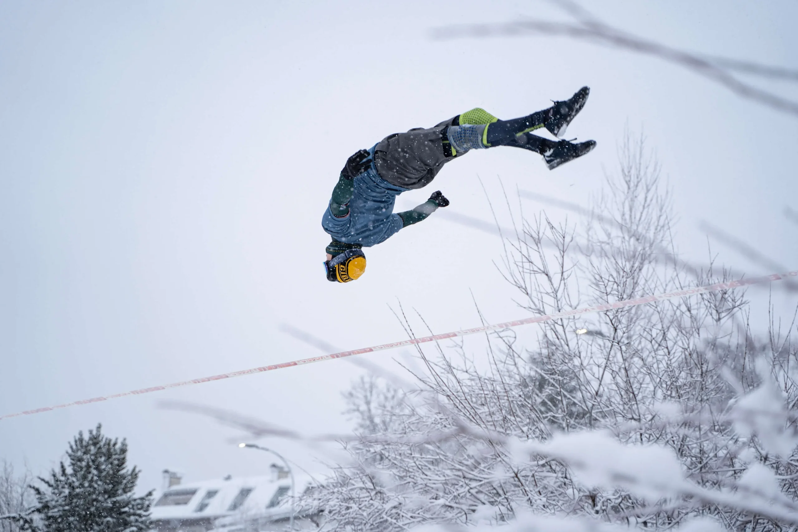 Eine Person in Winterkleidung und mit gelbem Helm vollführt einen Rückwärtssalto über eine schneebedeckte Slackline im Freien, wobei verschneite Bäume und Dächer eine malerische Kulisse bilden.