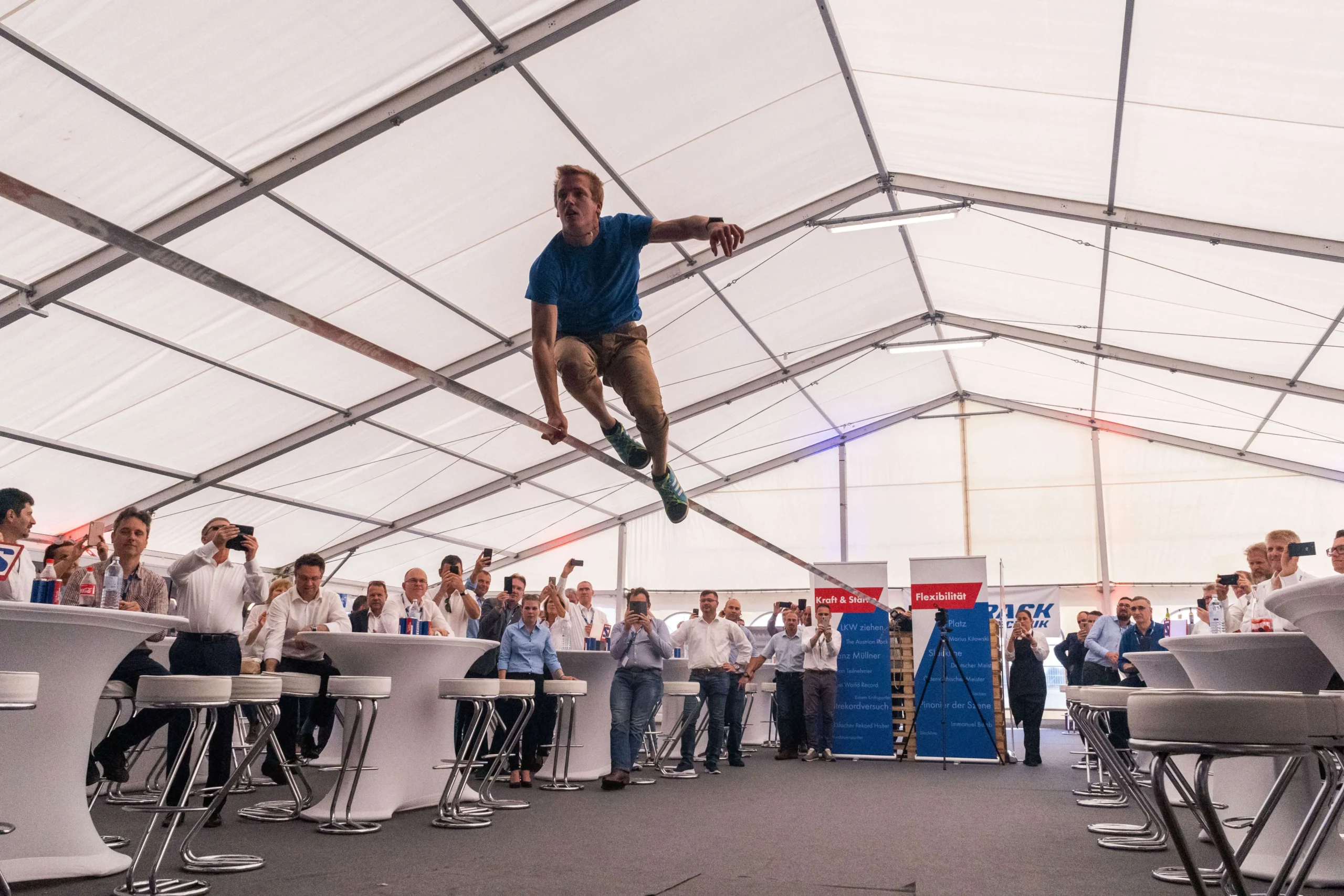 Ein Mann balanciert auf einer Slackline in einem großen Veranstaltungszelt in der Luft, umgeben von Zuschauern und Fotografen an Stehtischen.