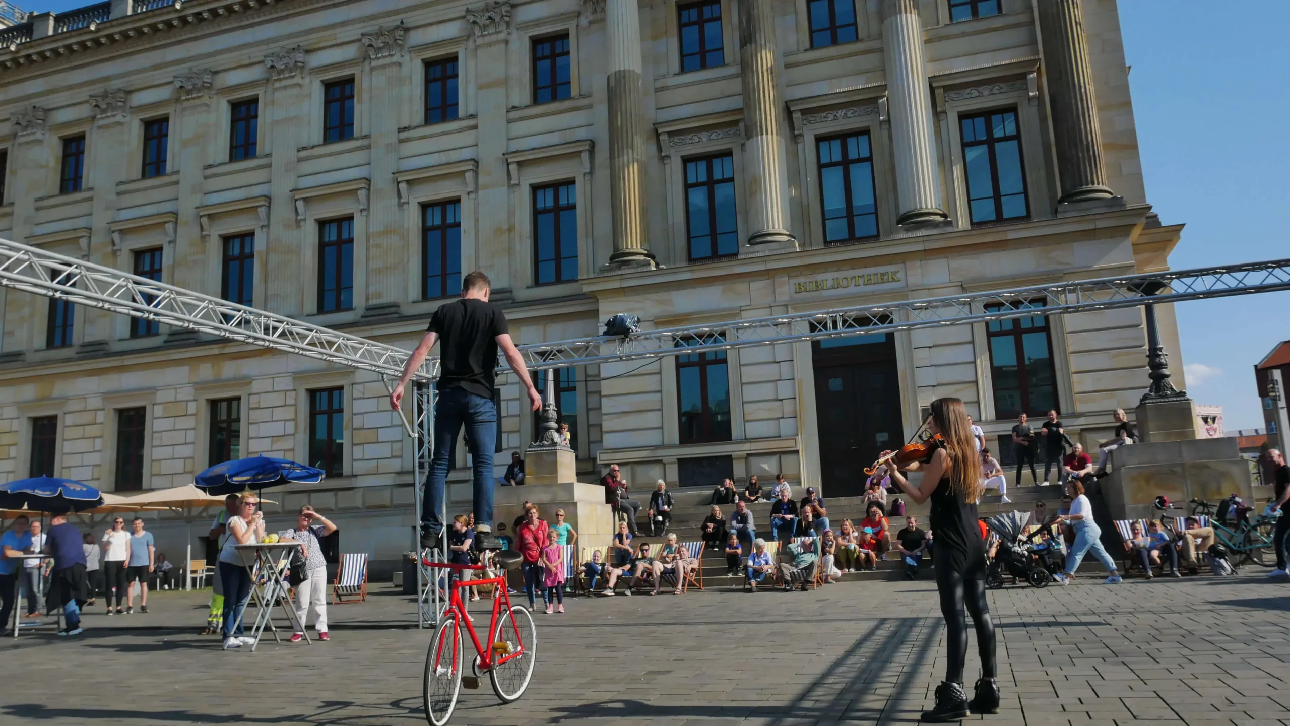 Ein Straßenkünstler fährt auf einem hohen Einrad, während eine andere Person vor einem großen Gebäude mit Säulen Geige spielt und damit eine Menschenmenge anzieht - ein einnehmender Moment des Stadtmarketings auf dem belebten Platz im Freien.