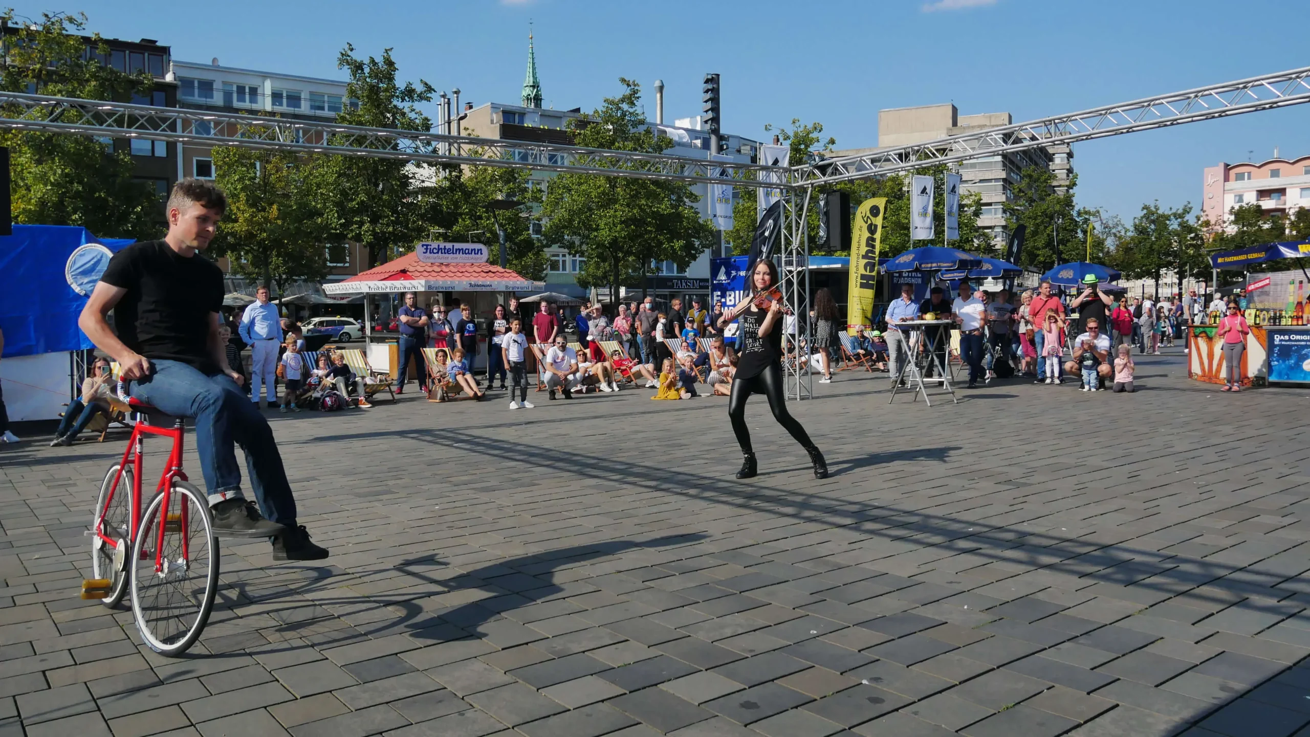 Ein junger Mann fährt auf einem Rad, während eine Frau mit einem Mikrofon vor ihm steht. Er tritt an einem sonnigen Tag auf einem Platz im Freien bei einer Stadtmarketing-Veranstaltung mit einem sitzenden Publikum und Zelten im Hintergrund auf.