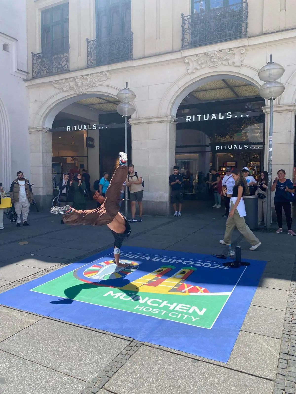 Ein Breakdancer macht einen einhändigen Handstand auf einer blauen Matte mit der UEFA EURO 2024 München Host City, als Teil des Stadtmarketings, vor einem Rituals-Geschäft, während eine Menschenmenge zusieht und Fotos macht.