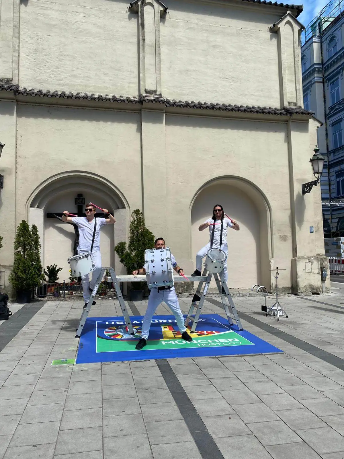 Drei weiß gekleidete Straßenkünstler spielen im Rahmen einer Stadtmarketing-Veranstaltung auf Trommeln, die an Leitern vor einem beigen Gebäude mit gewölbten Türöffnungen befestigt sind. Einer steht auf dem Boden, während die beiden anderen auf Leitern stehen.