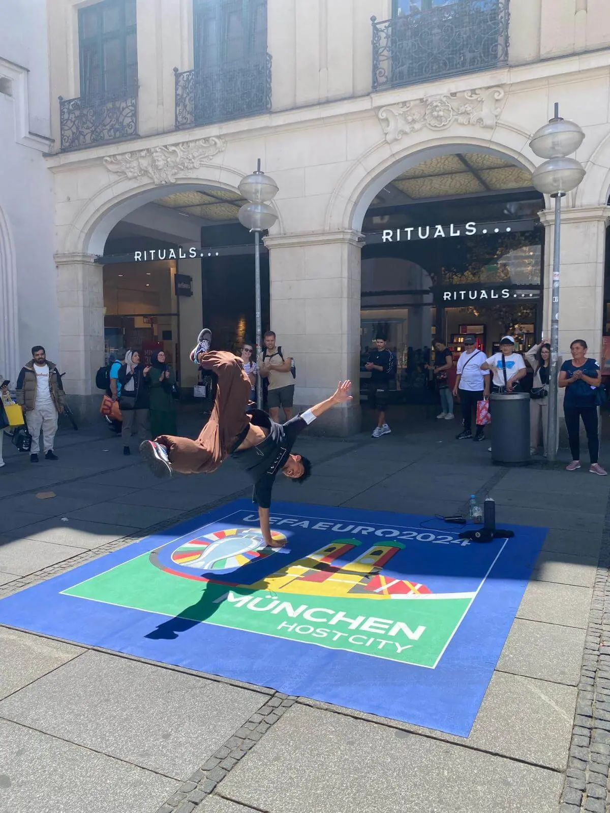 Ein Breakdancer macht einen einhändigen Handstand auf einer bunten Matte mit dem Branding der EURO 2024 München Host City und des Stadtmarketings vor einem Gebäude mit Bogenfenstern und einem RITUALS-Schild, während die Zuschauer zusehen.