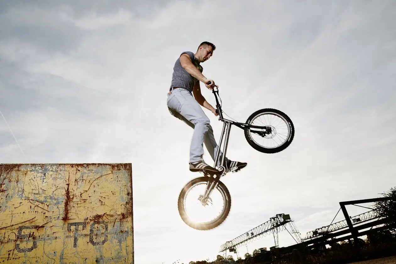 Eine Person führt während einer Trial Bike Show auf einem BMX-Rad in der Nähe einer mit Graffiti bedeckten Rampe einen Hochsprungtrick aus, wobei sich die Silhouette gegen den Himmel abhebt und im Hintergrund Industrieanlagen zu sehen sind.