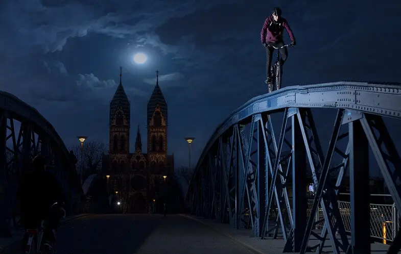 Eine Person vollführt eine waghalsige Trial Bike Show, indem sie nachts mit einem Fahrrad am oberen Geländer einer Metallbrücke entlangfährt. Der Vollmond und eine Kathedrale mit Zwillingstürmen beleuchten die Szene, während ein anderer Radfahrer die Brücke unter ihm überquert.