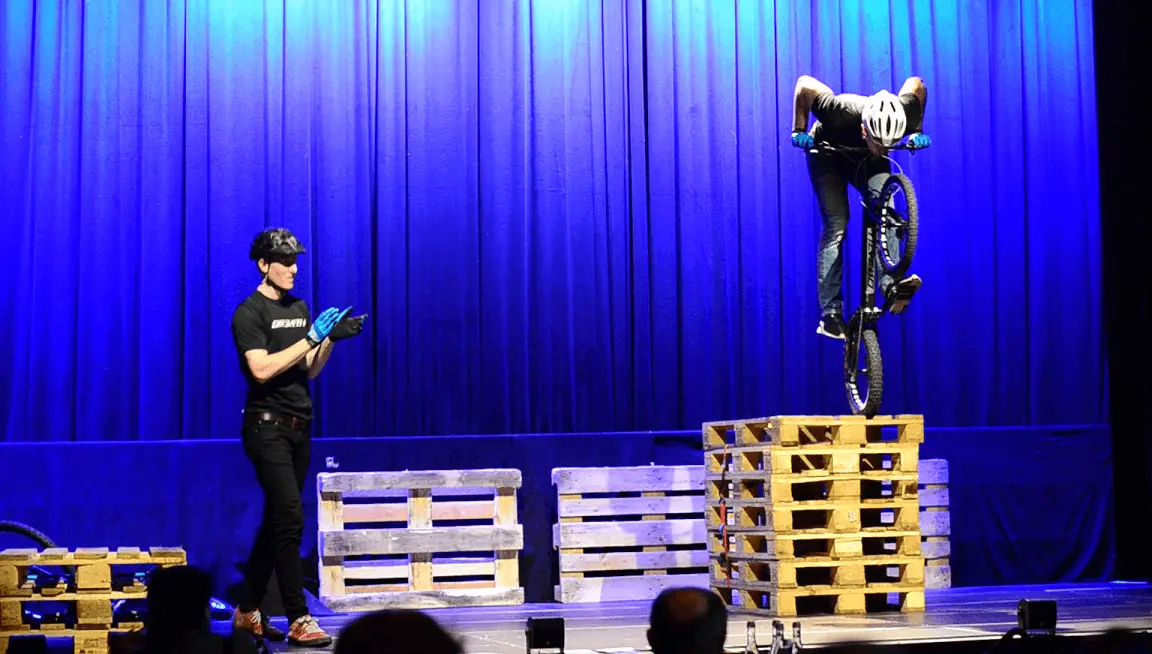 Während einer Trial Bike Show balanciert ein Radfahrer auf gestapelten Holzpaletten, während eine andere Person in Handschuhen und mit Helm in der Nähe steht und klatscht. Blaue Scheinwerfer beleuchten die Bühne, die von dunklen Vorhängen umrahmt ist.
