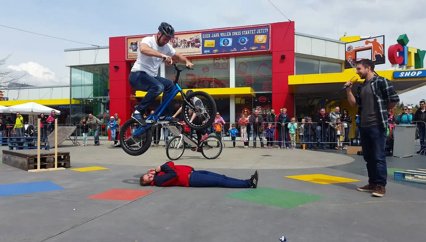 Während einer aufregenden Trial Bike Show springt ein Mann auf einem BMX-Rad über eine am Boden liegende Person, während eine Menschenmenge hinter einer Absperrung zuschaut und ein buntes Gebäude eine lebendige Kulisse bildet. Ein anderer Mann steht in der Nähe und hält ein Mikrofon.