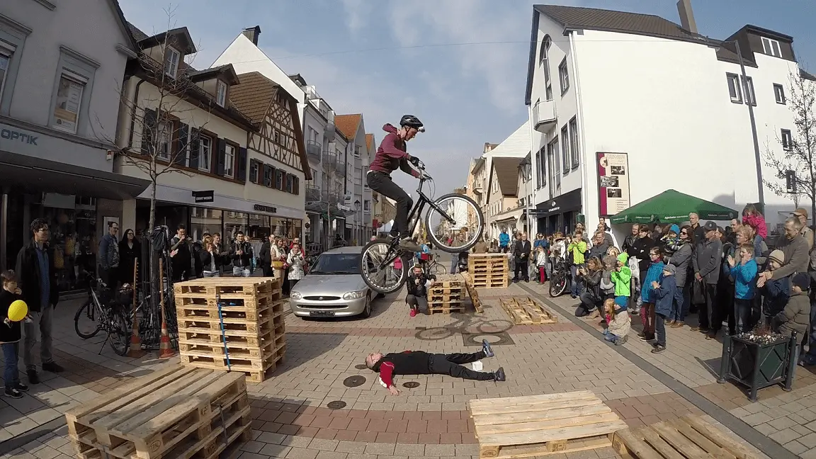 Während einer Trial Bike Show vollführt ein Radfahrer einen waghalsigen Stunt auf gestapelten Holzpaletten und springt mit seinem Fahrrad über eine Person, die auf dem Bürgersteig liegt, während eine begeisterte Menge auf einer Stadtstraße zusieht.