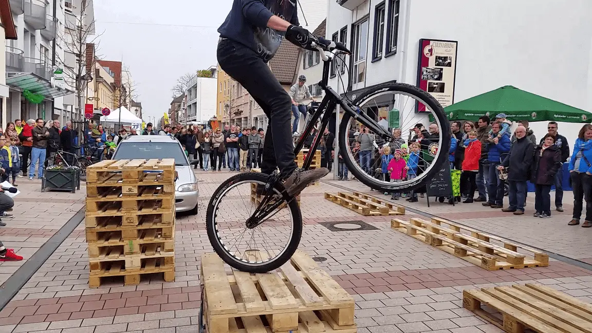 Ein Radfahrer vollführt einen waghalsigen Trick, indem er während einer Trial Bike Show auf gestapelten Holzpaletten auf dem Hinterrad balanciert, während eine Menschenmenge in einer städtischen Umgebung zusieht.