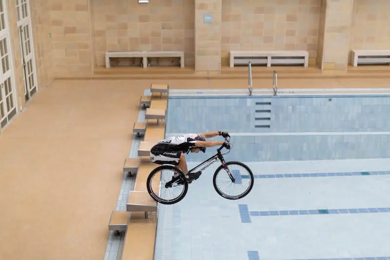 Eine Person, die einen Trial-Bike-Show-Stunt vorführt, bei dem sie mit einem Fahrrad vom Rand eines Sprungbretts in ein leeres Hallenbad stürzt.