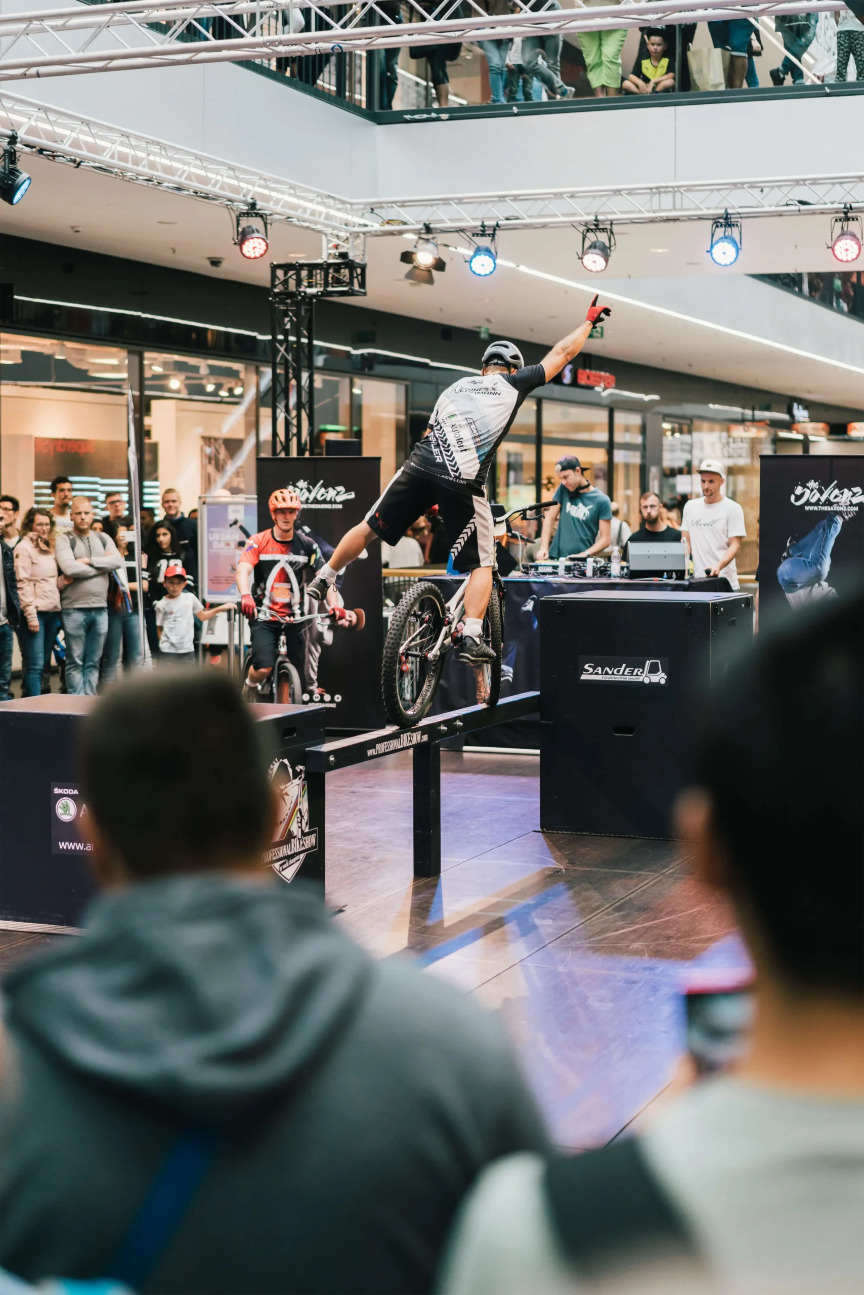 Ein Radfahrer führt während einer aufregenden Trial Bike Show auf einem schmalen Balken einen Balanciertrick auf einem Fahrrad aus, während eine Menschenmenge in einem Einkaufszentrum zusieht und Fotos macht.