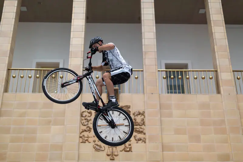 Ein Radfahrer mit Helm und Sportkleidung vollführt einen Sprung auf einem Mountainbike während einer Trial Bike Show vor einem beigen Gebäude mit Säulen und einem Uhrenemblem an der Wand.