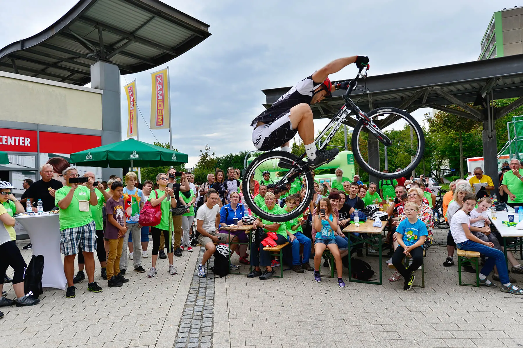 Ein Radfahrer vollführt einen hohen Sprung während einer spannenden Trial Bike Show vor einer großen, jubelnden Menge bei einer Veranstaltung im Freien mit Tischen, grünen Regenschirmen und Gebäuden im Hintergrund.